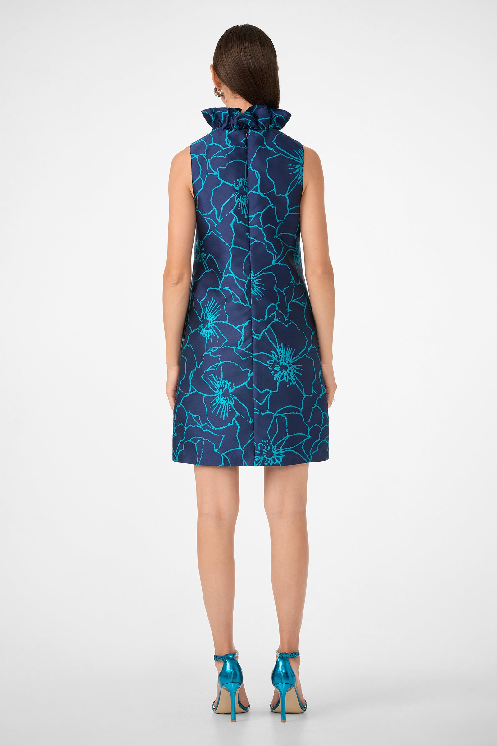 A woman with straight brown hair pulled back wears the Edina Dress, a sleeveless, knee-length blue dress with a bold floral print and a ruffled high collar, paired with teal high-heeled sandals.