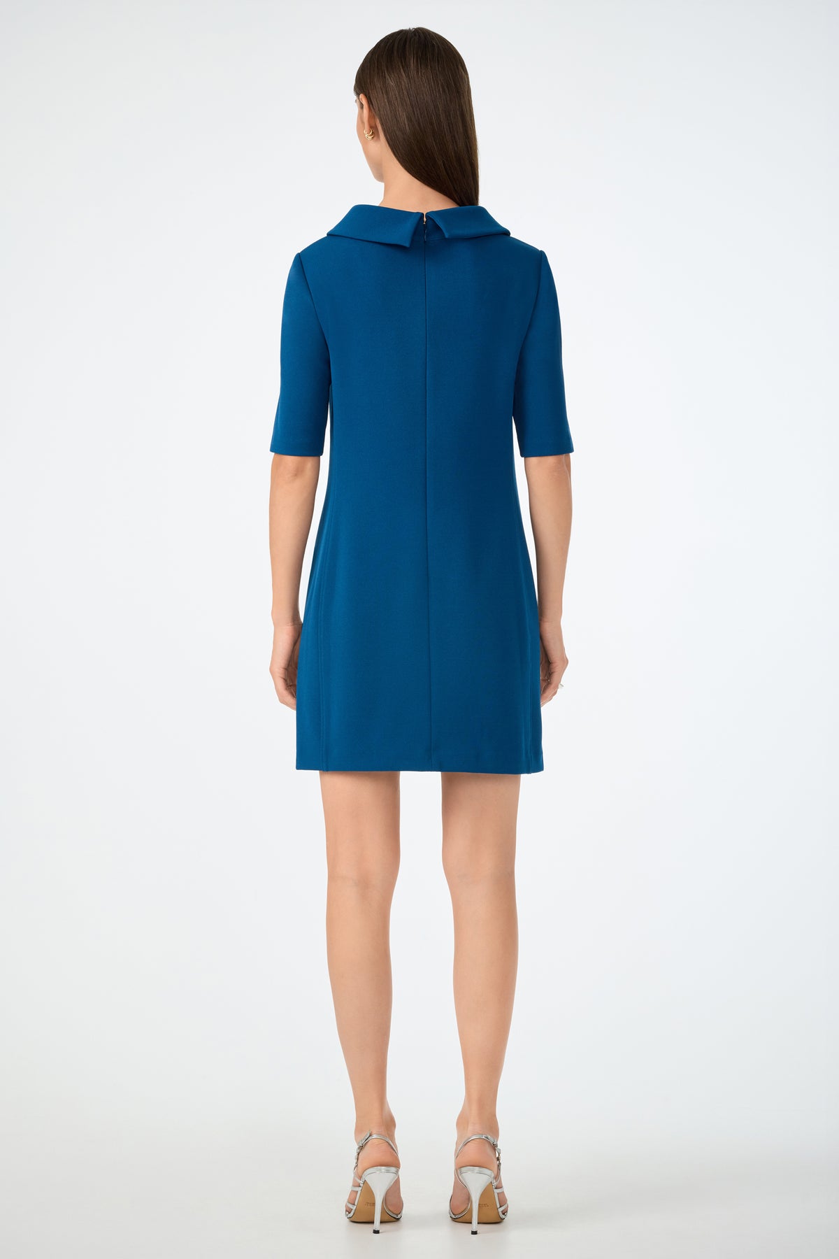 A woman with straight brown hair faces away, modeling the Maleko Dress—a teal, knee-length style with short sleeves and a wide collar—paired with strappy silver heels against a plain white background.