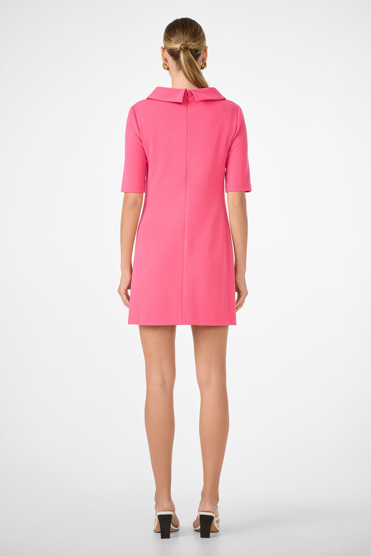 A woman with her hair tied back wears the Maleko Dress—a collared, short-sleeved, knee-length bright pink dress—paired with open-toe heels. She stands facing away against a plain white background.