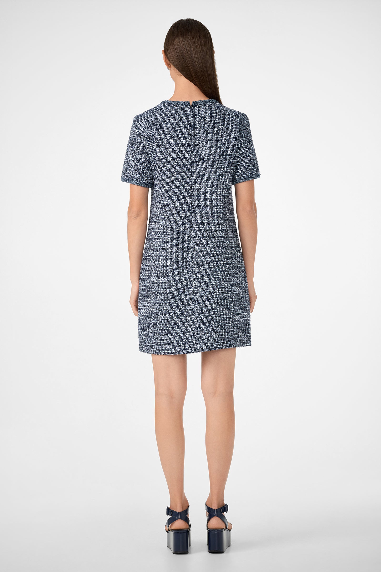 A woman with straight brown hair, seen from behind, wears the Akara Dress—a short-sleeve, textured blue dress—with dark blue platform sandals against a plain white background.