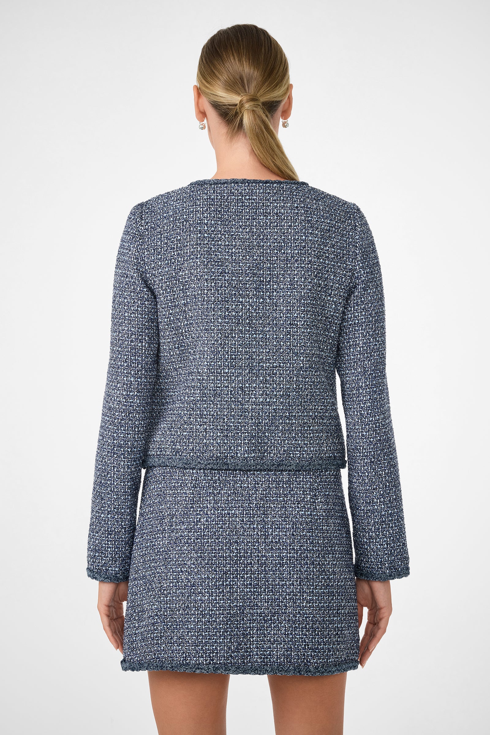A woman with blonde hair in a low ponytail is shown from the back wearing the Kiui Blazer, a textured blue and white long-sleeve jacket with a matching short skirt, against a plain white background.