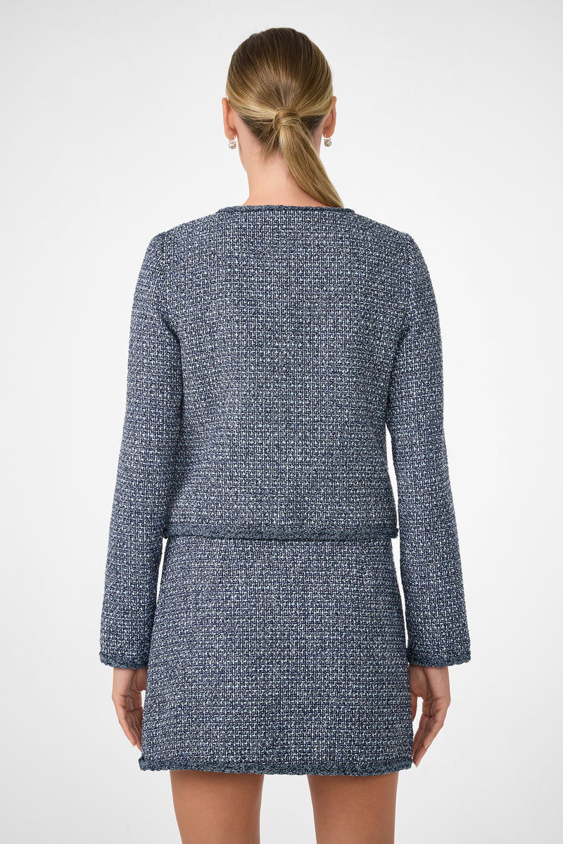 A woman with blonde hair in a low ponytail is shown from the back wearing the Kiui Blazer, a textured blue and white long-sleeve jacket with a matching short skirt, against a plain white background.