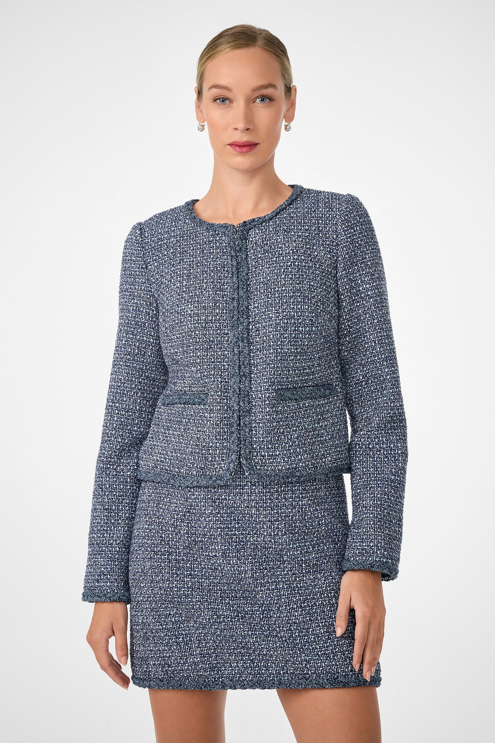 A woman confidently poses against a plain white background, wearing the Kiui Blazer—a tailored blue and white tweed jacket with a matching mini skirt.