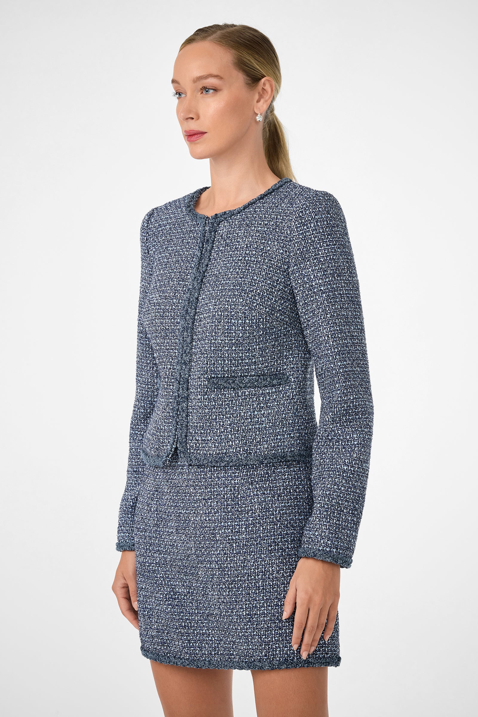A woman stands against a plain background wearing the Kiui Blazer, a blue and white textured tweed jacket, with matching skirt. Her hair is pulled back as she gazes slightly to the side with a neutral expression.