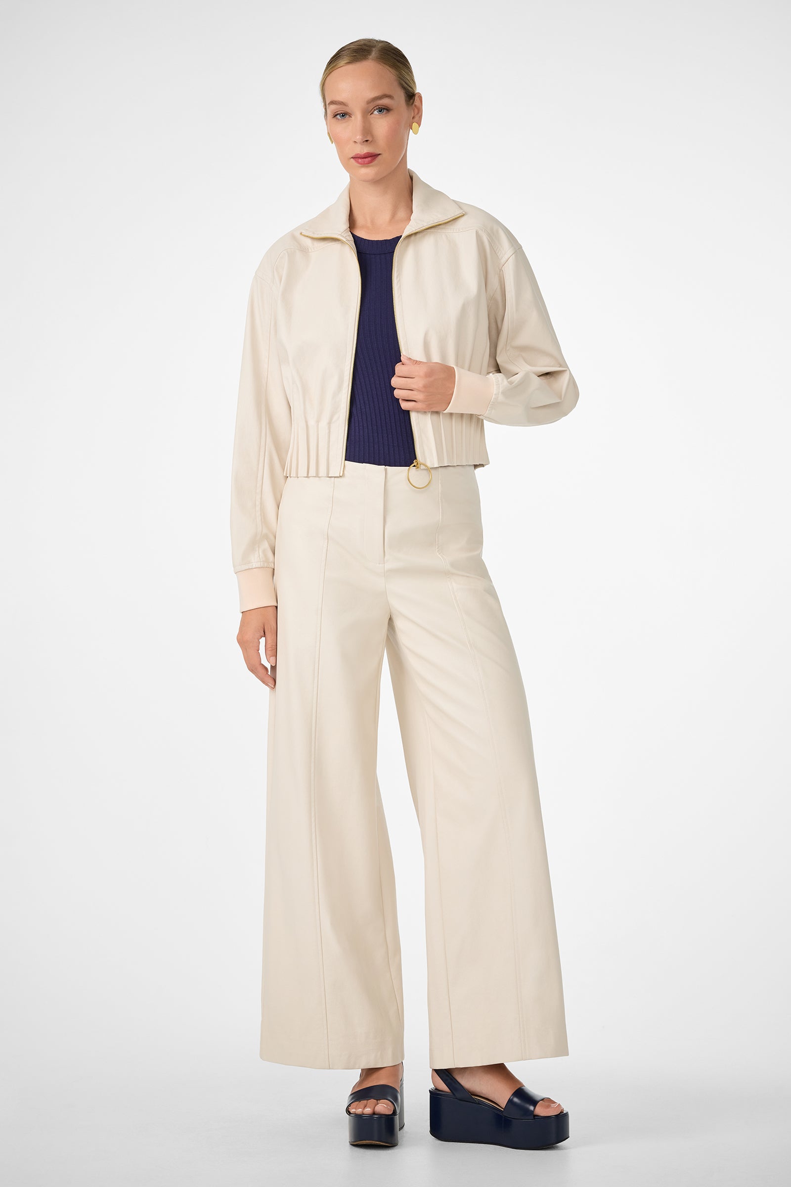 A woman with her hair pulled back and a neutral expression stands against a plain background, wearing the cream-colored Outrun Jacket, matching wide-leg high-waisted pants, a navy top, and navy platform sandals.
