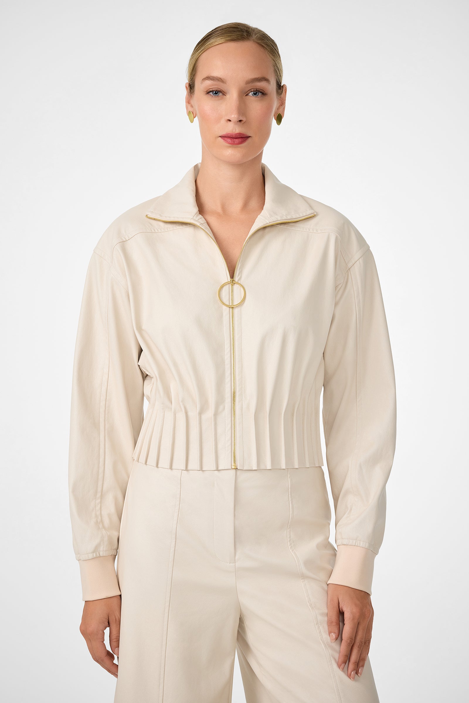 A woman wearing the Outrun Jacket—a light beige collared jacket with a gold ring zipper and pleated waist—paired with matching high-waisted pants, stands against a plain white background.