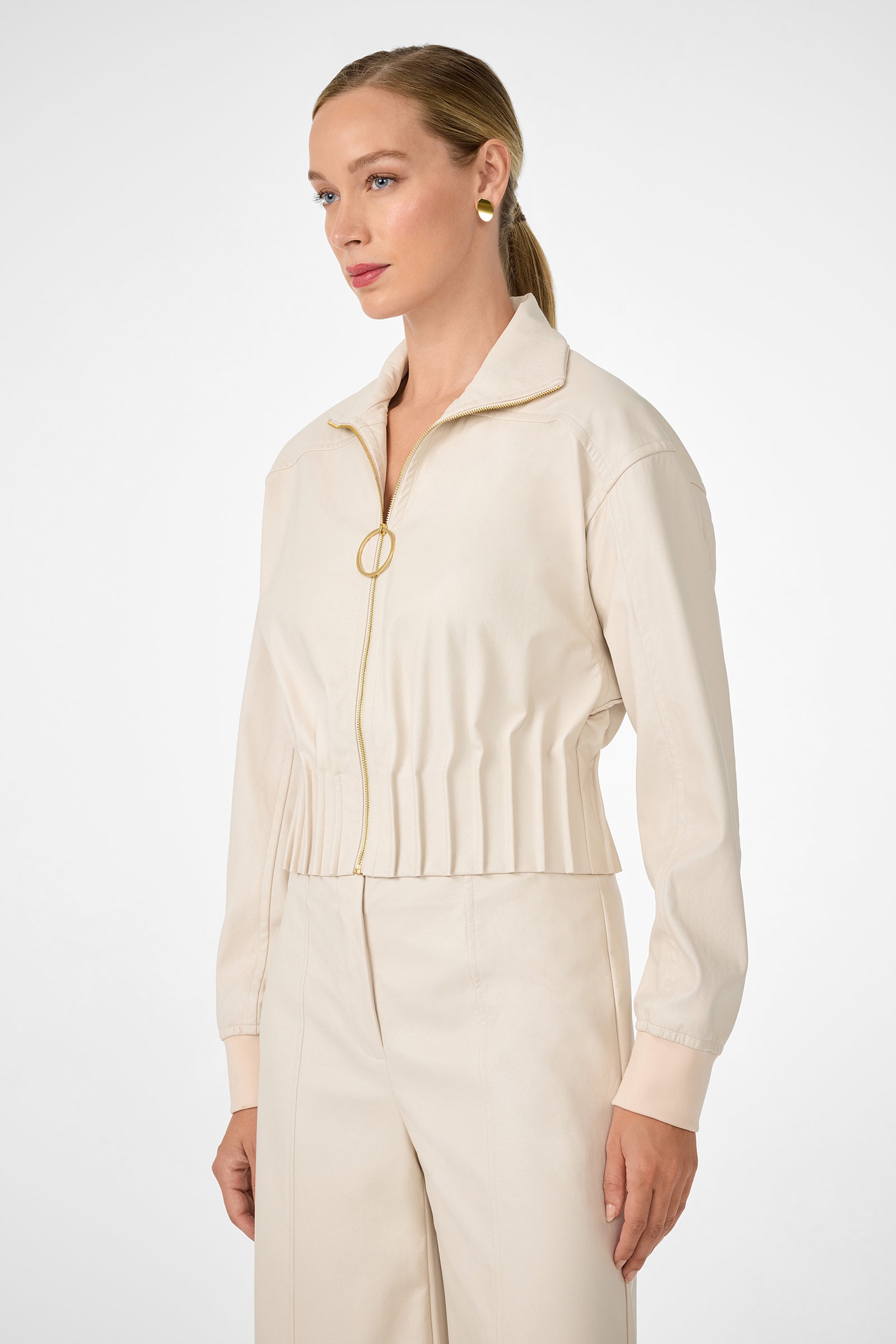 A woman with blonde hair in a low ponytail wears the cream Outrun Jacket with matching high-waisted pants, standing against a plain white background and looking to the side.