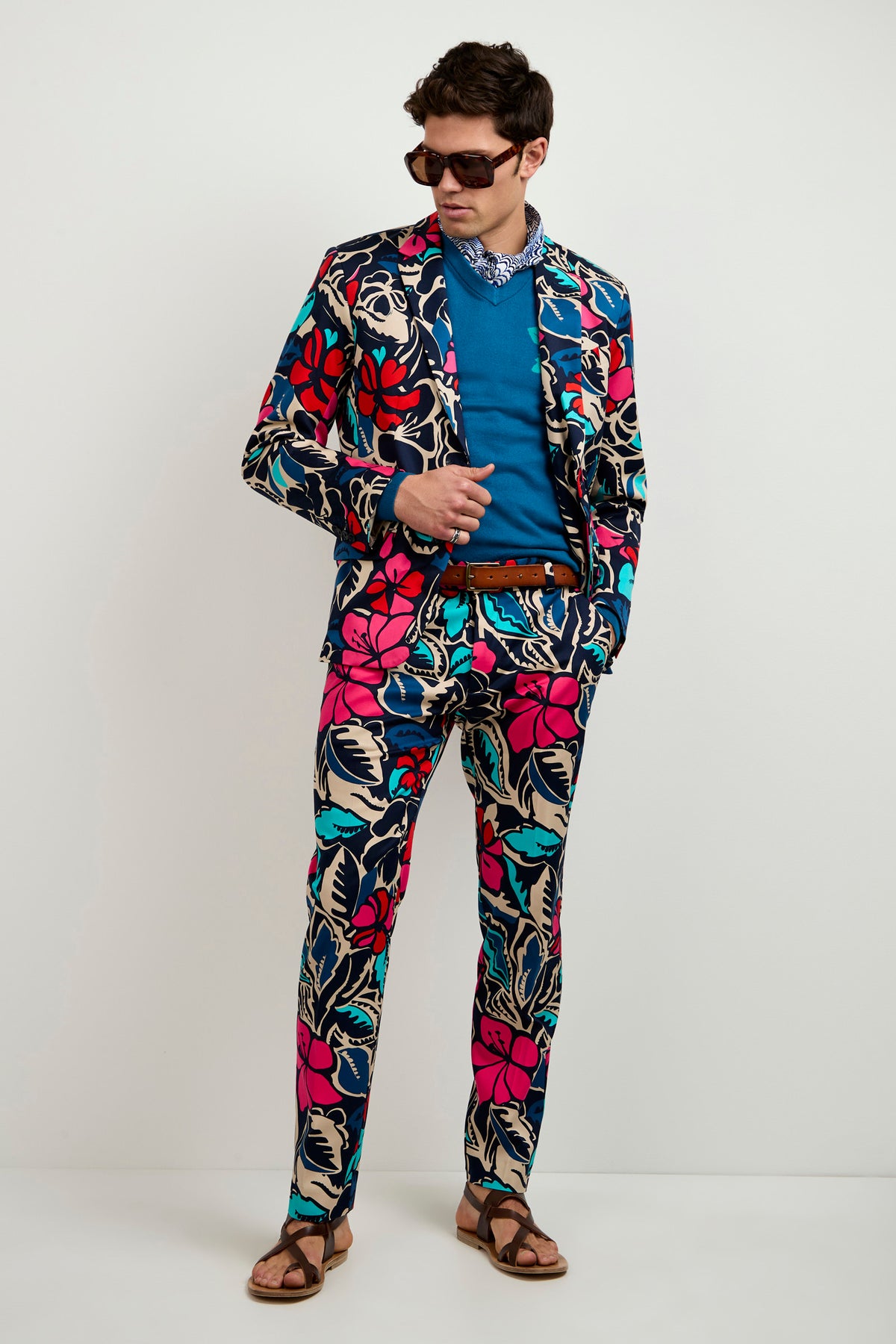 A man in the Thurston Blazer - Copacabana, paired with a blue shirt, sunglasses, neck bandana, brown belt, and sandals stands against a plain light background, looking down at his outfit.