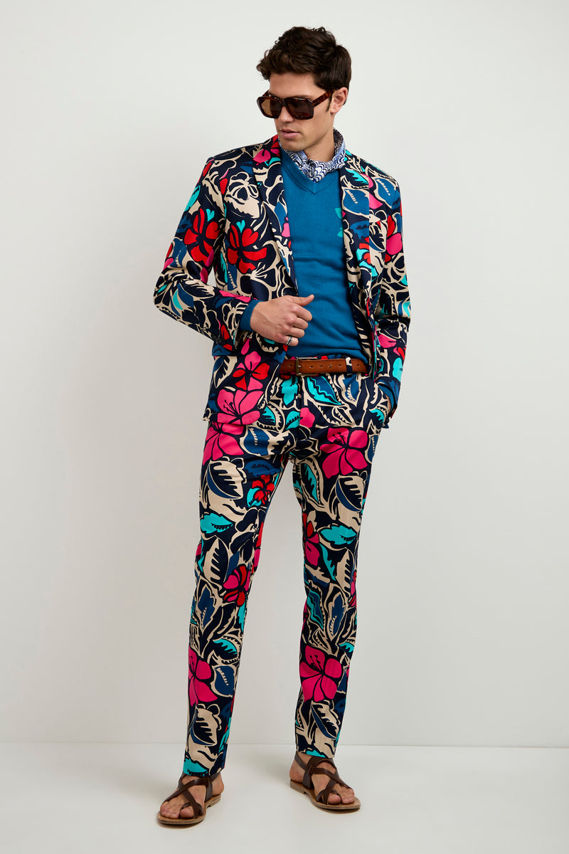 A man in the Thurston Blazer - Copacabana, paired with a blue shirt, sunglasses, neck bandana, brown belt, and sandals stands against a plain light background, looking down at his outfit.