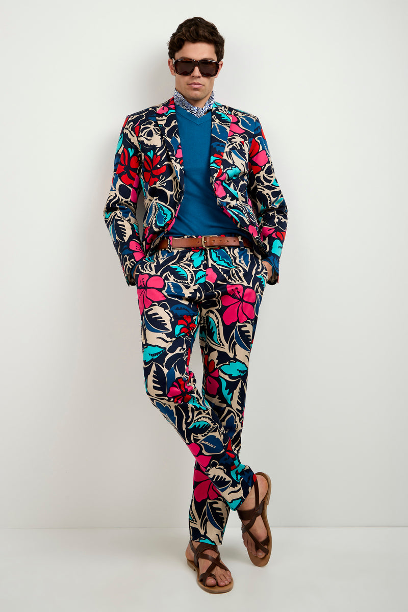 A man wearing the Clyde Slim Trouser - Copacabana, paired with a blue shirt, brown belt, patterned scarf, sandals, and sunglasses stands against a white wall with his hands in his pockets and one foot crossed over the other.