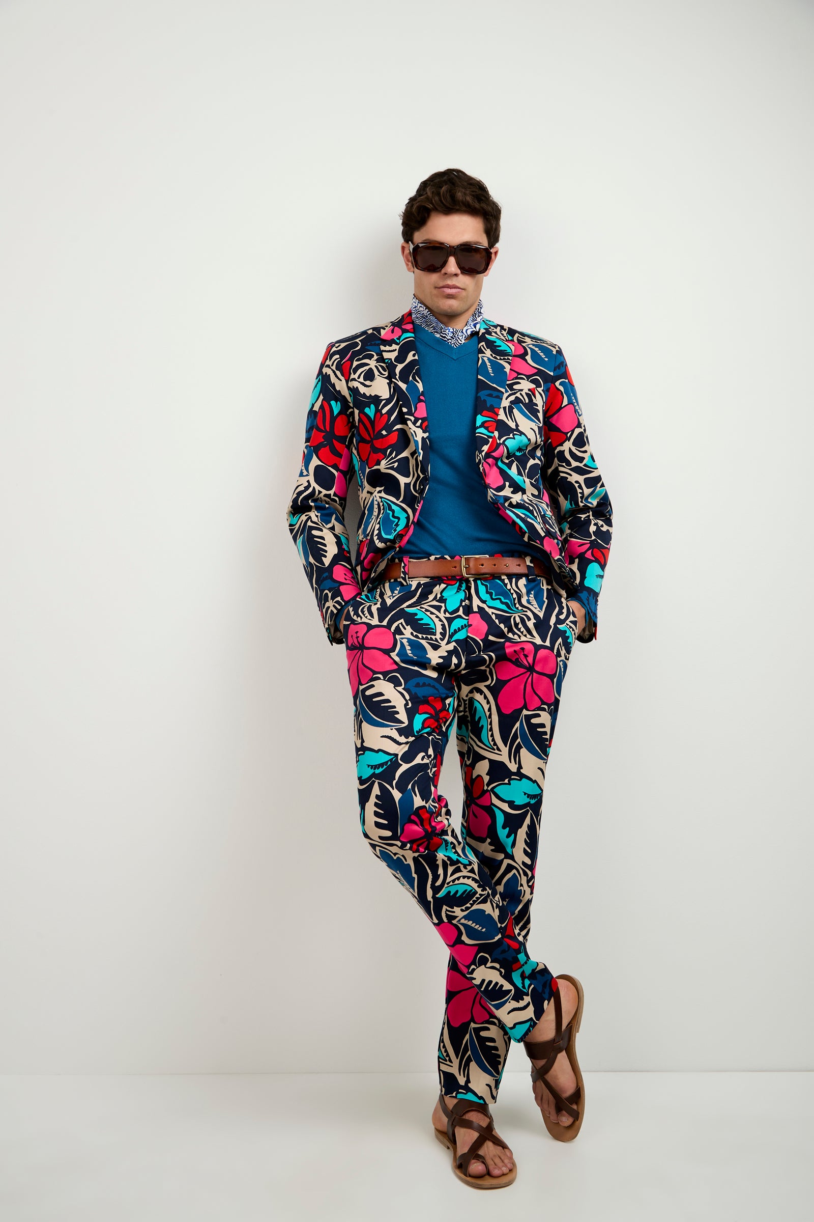 A man confidently stands against a plain white wall, wearing the Thurston Blazer - Copacabana, featuring vibrant floral patterns in red, pink, blue, and beige, paired with a blue shirt, brown belt, sandals, and dark sunglasses.
