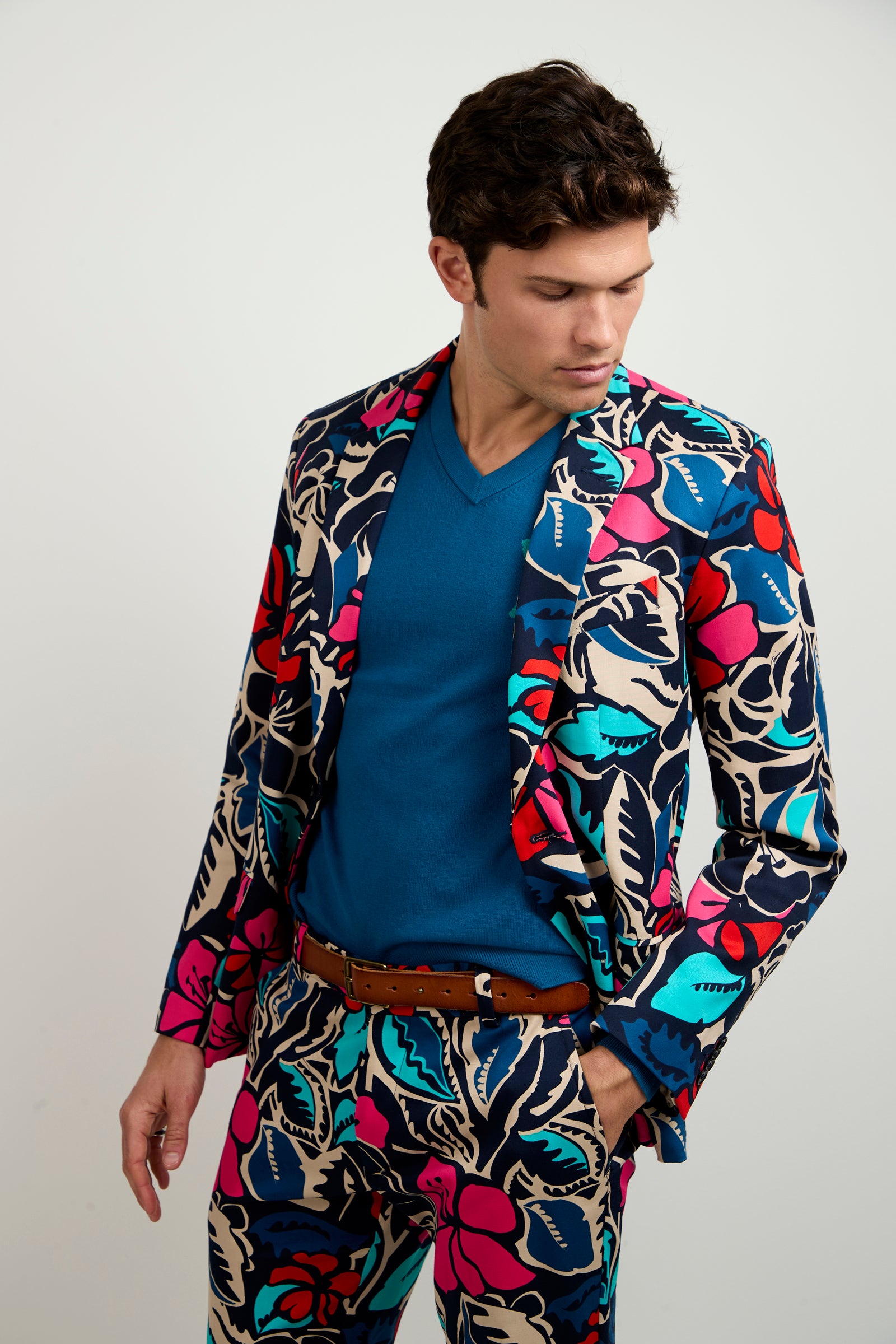 A man wears the Thurston Blazer - Copacabana, featuring a bold, colorful floral pattern over a blue V-neck shirt, with one hand in his pocket against a plain light background.