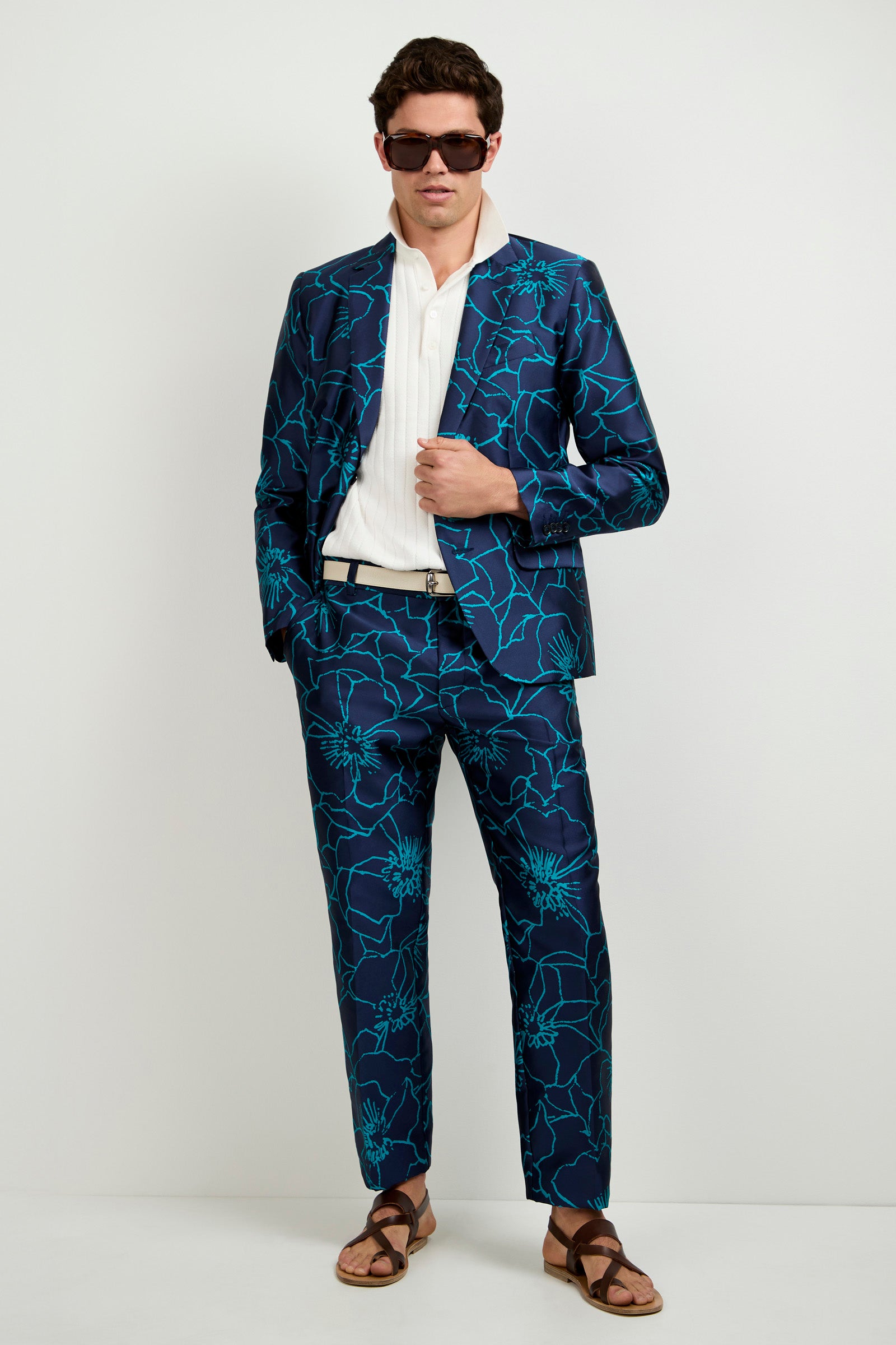 A man wears the Clyde Slim Trouser - Jucara Jacquard with a dark blue suit featuring turquoise floral patterns, sunglasses, a white shirt, black belt, and brown sandals, standing against a plain light background with one hand in his pocket.