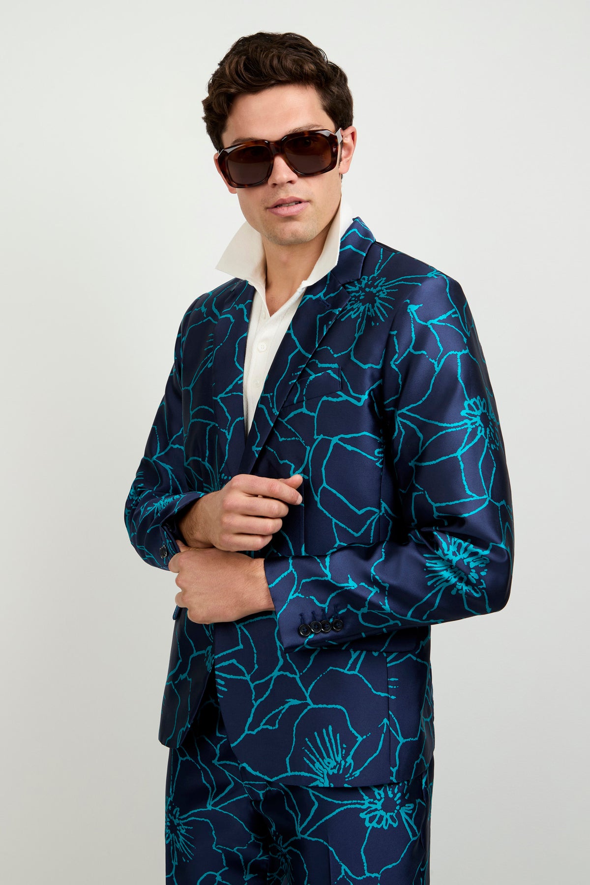 A man wearing oversized sunglasses and the Thurston Blazer - Jucara Jacquard, featuring bold turquoise floral patterns over a navy blue base, stands against a plain background, adjusting his jacket over a crisp white shirt.