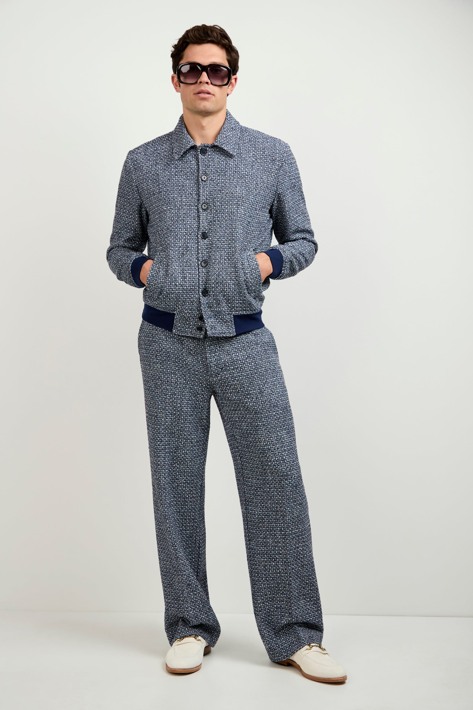 A man stands against a plain background wearing Buckingham Tweed Trousers with a matching blue textured button-up jacket, white sneakers, and dark sunglasses, hands in pockets.