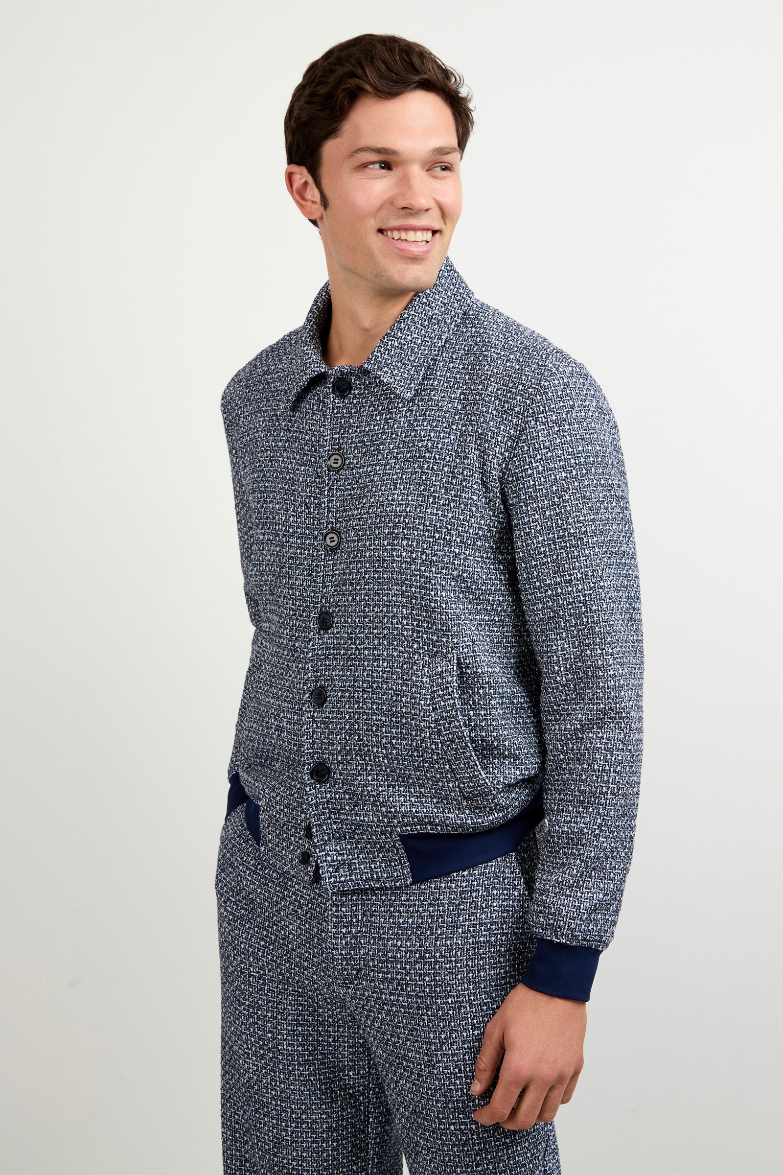 A man wears the Lowen Tweed Jacket, a textured blue and white button-up, with matching pants, standing against a plain light background and smiling as he looks to his left.
