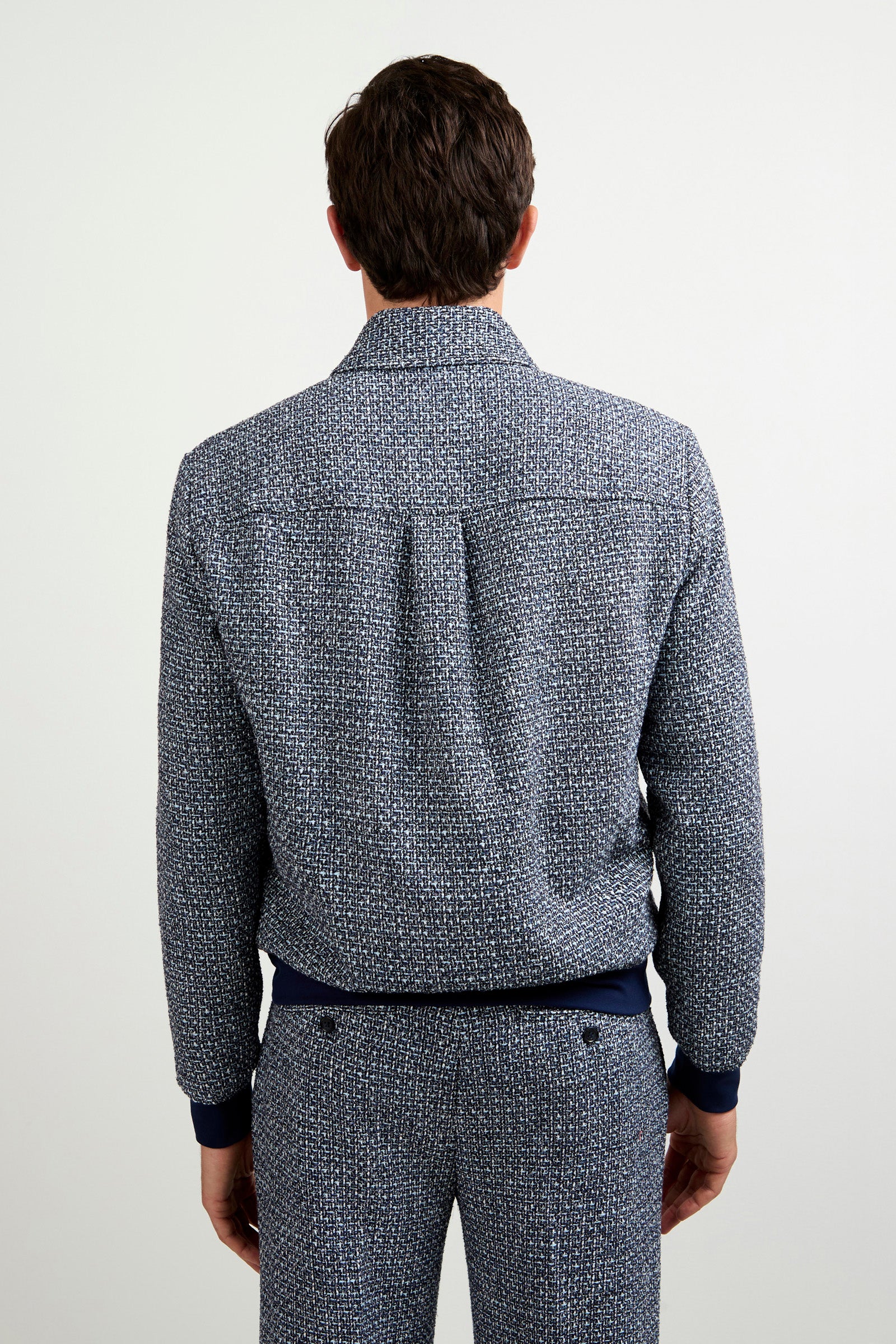 A man stands facing away from the camera, wearing the Lowen Tweed Jacket with matching blue and white patterned pants. The background is plain and light-colored.