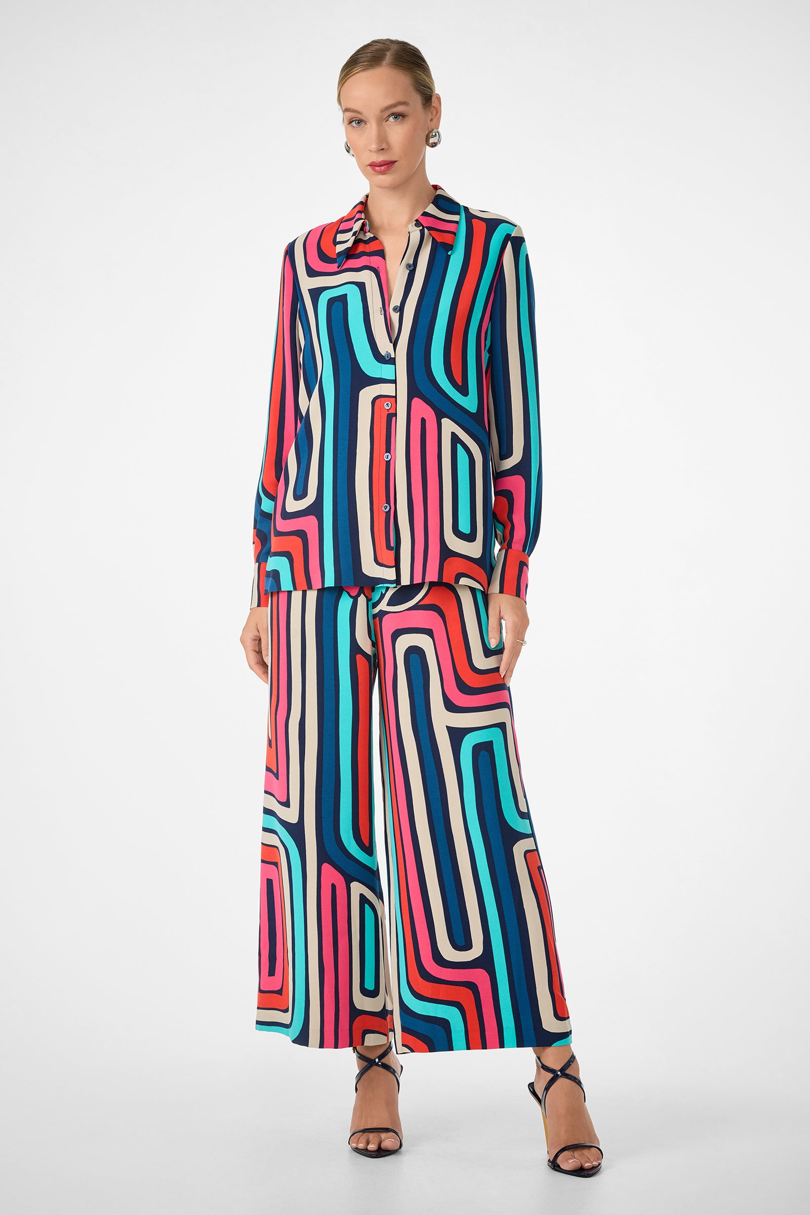 A woman wears the Montero Top, a long-sleeve button-up shirt featuring a bold geometric pattern in red, blue, pink, and black, paired with matching wide-leg pants and black strappy heels. Her hair is styled back.