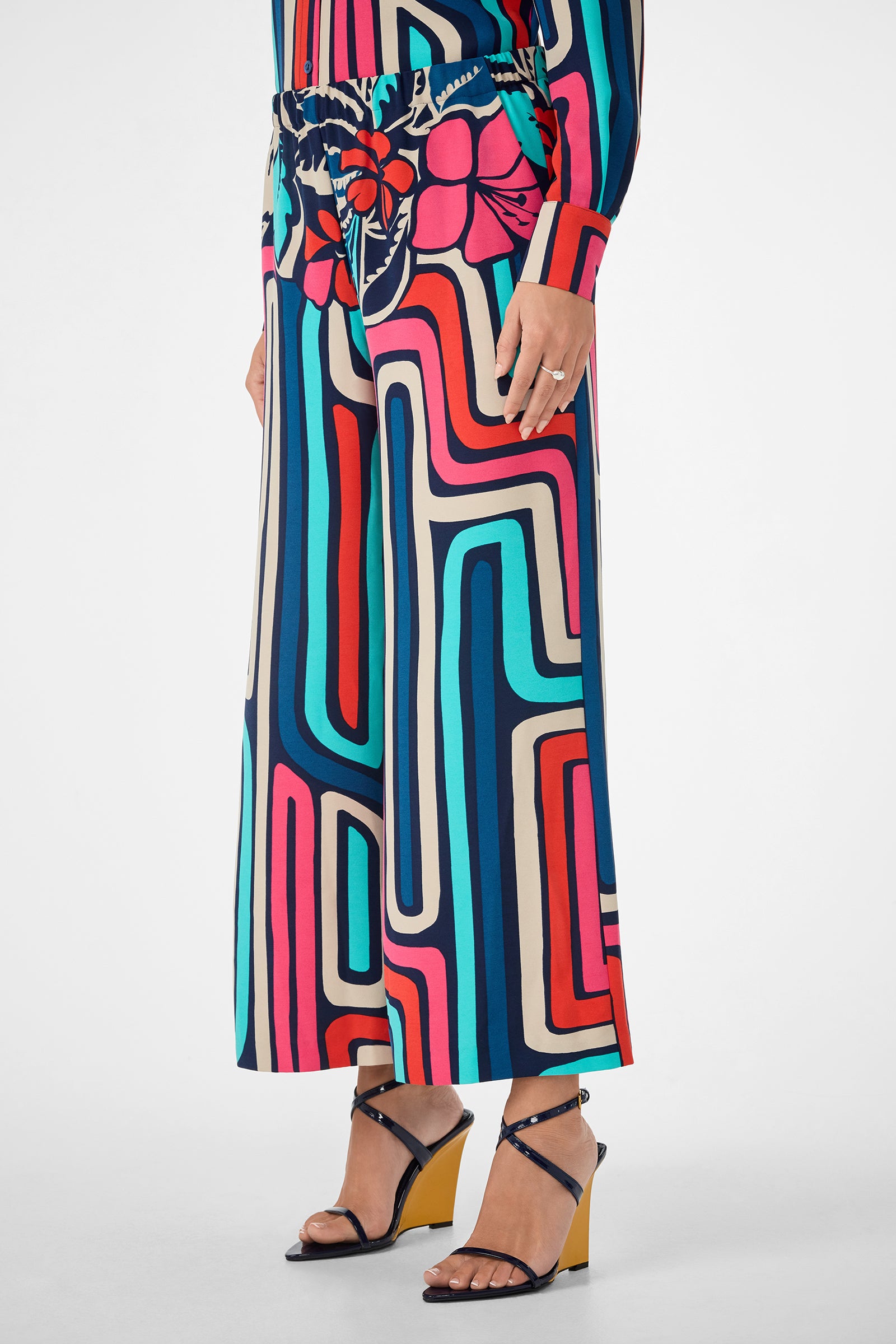 Only the lower half of the body is visible, showing a person in the Cubism Pant—wide-leg pants with a bold, colorful geometric and floral print—paired with a matching top and black strappy wedge sandals with yellow heels.