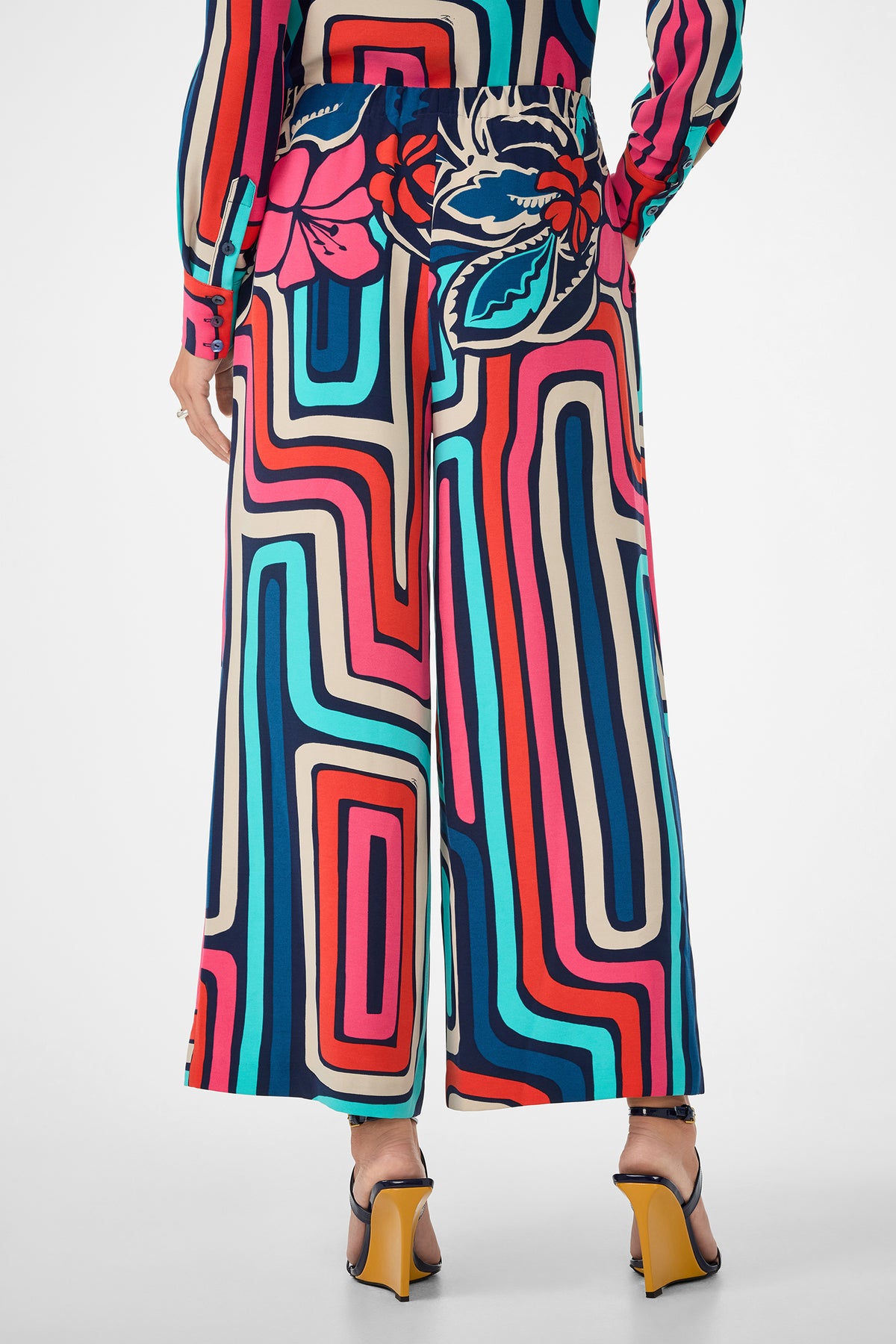 A person wearing the Cubism Pant, featuring a bold, colorful geometric and floral print in a wide-leg style, paired with high-heeled sandals. The image shows the back view from waist down.