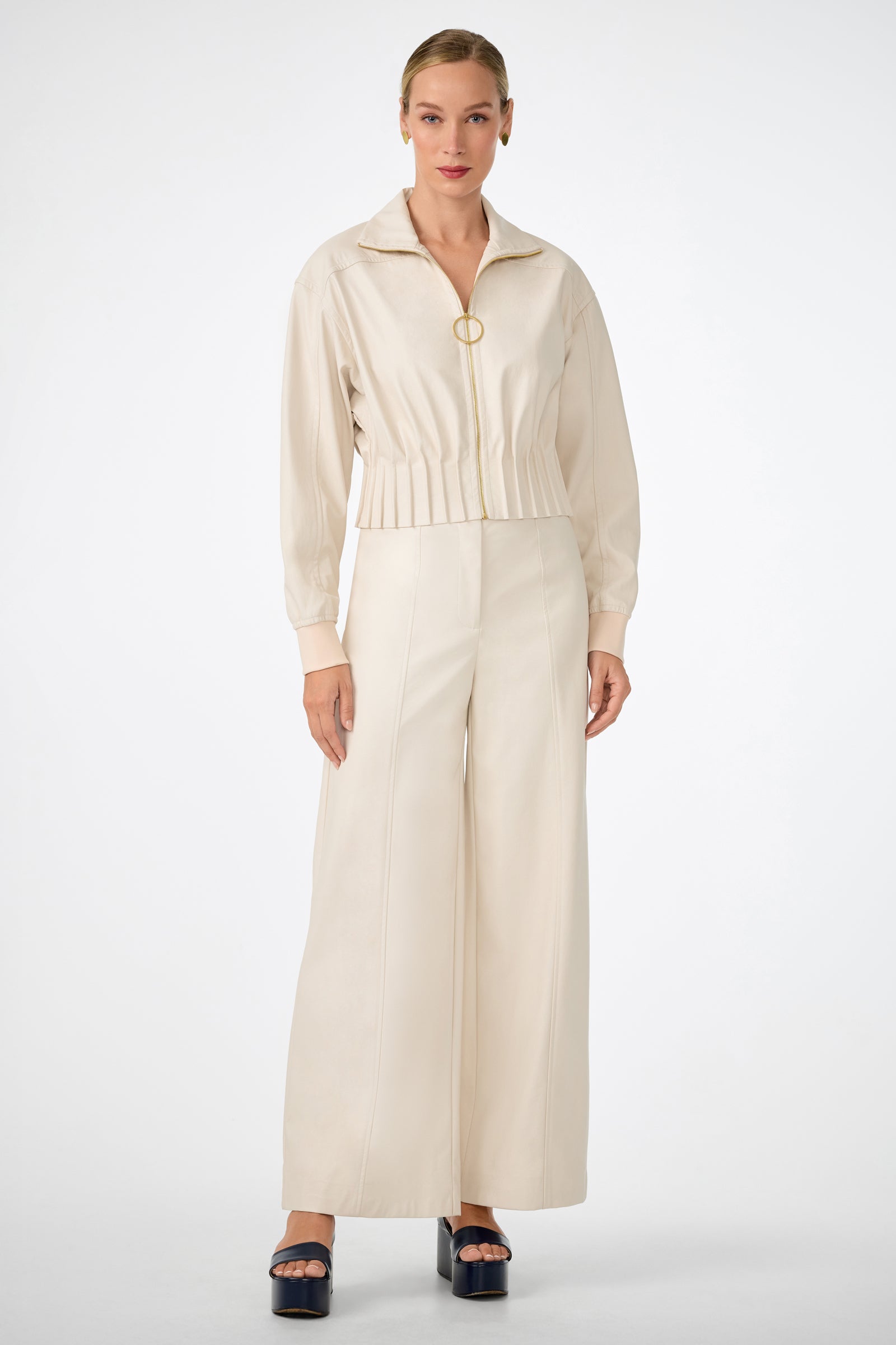 A woman stands facing forward in cream-colored high-waisted Parque Pants with a matching zip-up jacket and dark platform sandals, set against a plain white background.