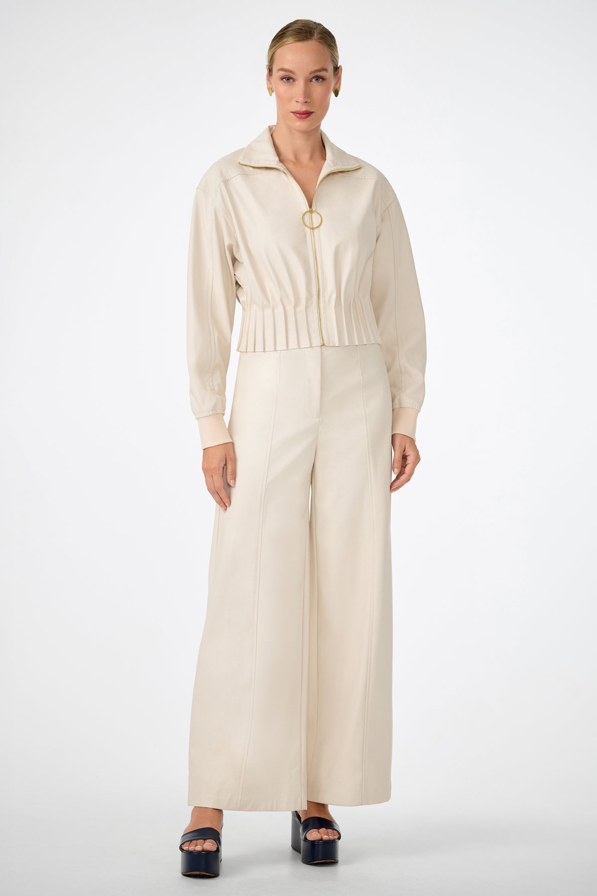 A woman stands facing forward in cream-colored high-waisted Parque Pants with a matching zip-up jacket and dark platform sandals, set against a plain white background.