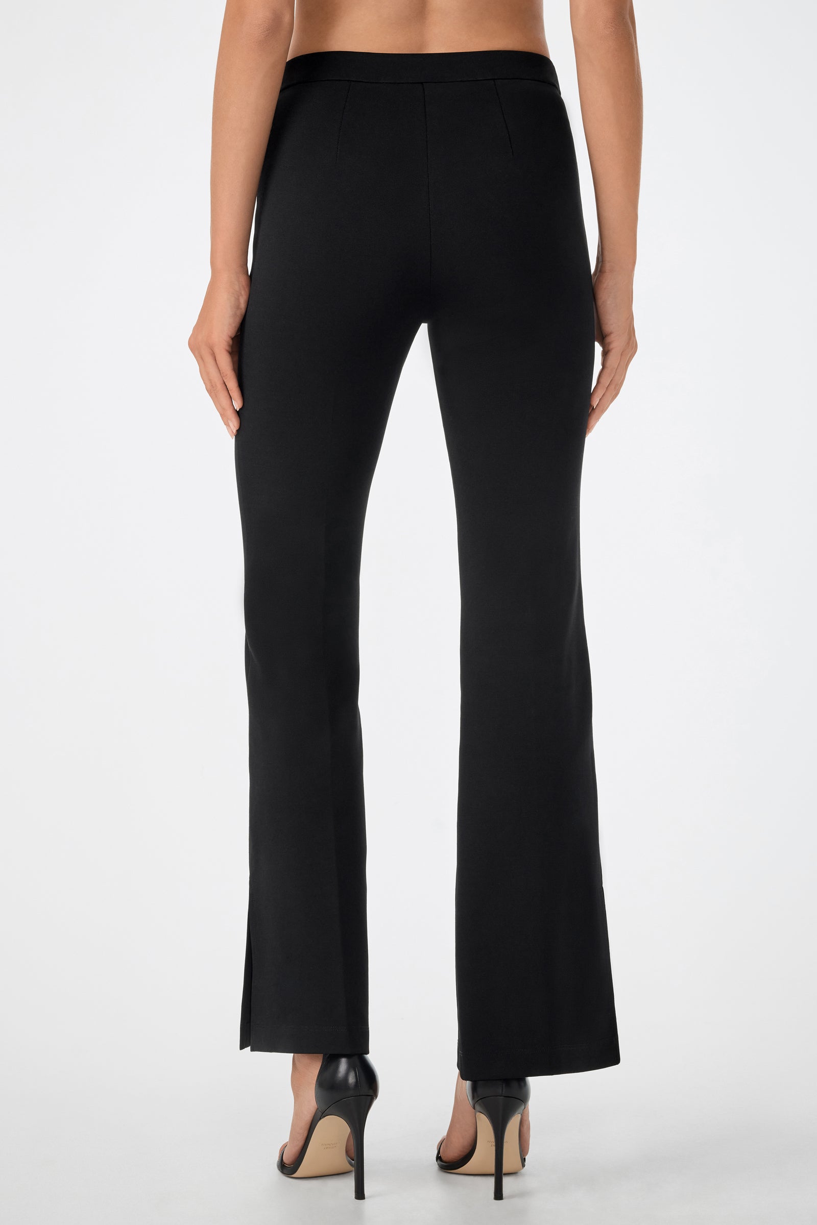 A woman is seen from the back wearing the Mona Pant—black, high-waisted, straight-leg pants with small hem slits—and black heeled sandals against a plain white background.