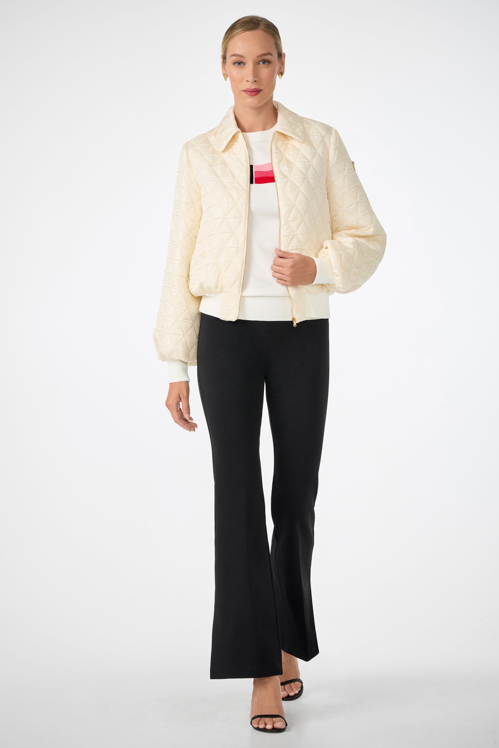 A woman stands against a white background wearing black Mona Pant flares, a cream quilted jacket, a white sweater with red and black stripes, and black open-toe heels.