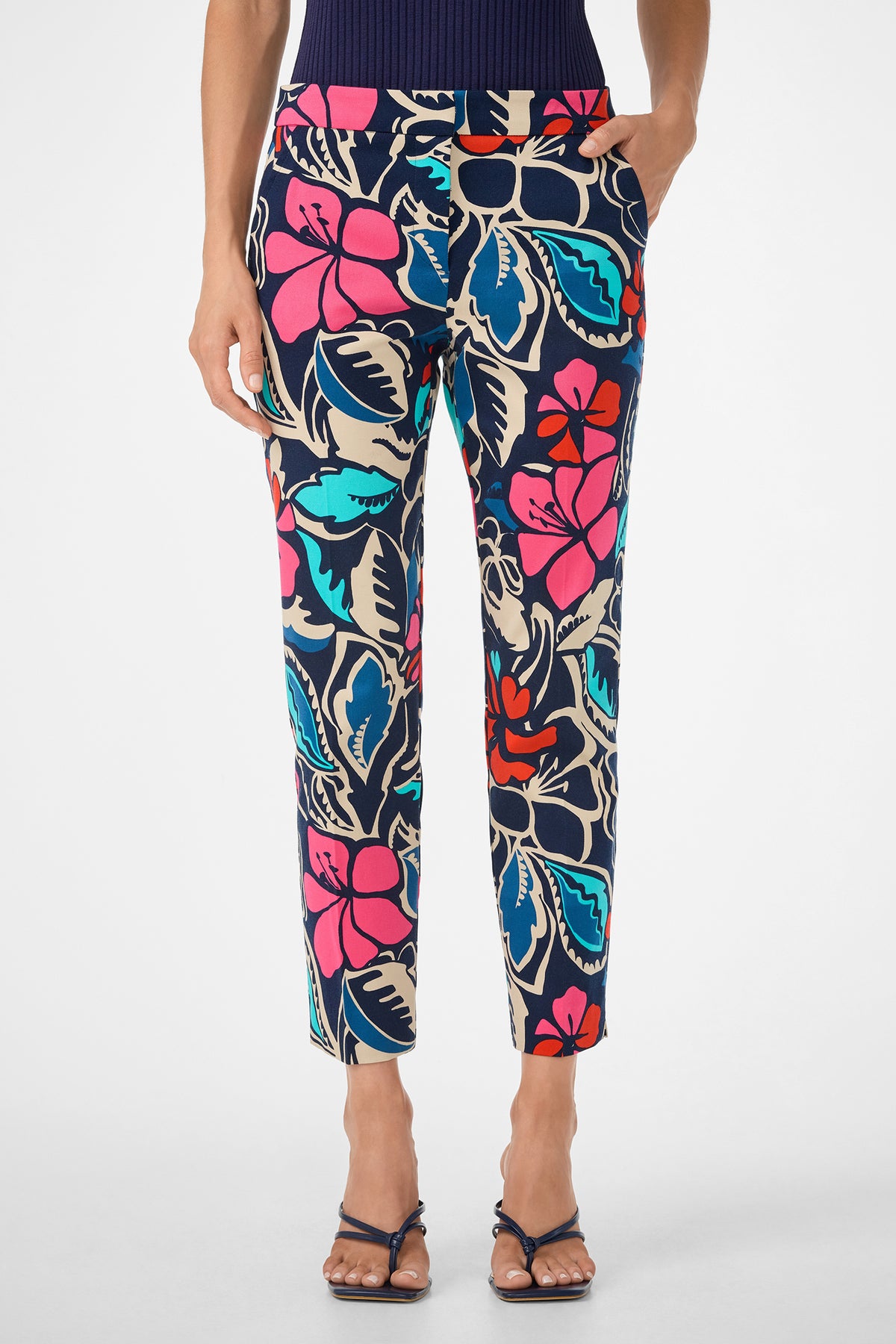 A person wearing Moss 2 Pant featuring colorful tropical floral prints in blue, pink, red, and cream, paired with strappy sandals and a dark top, hands in pockets against a white background.