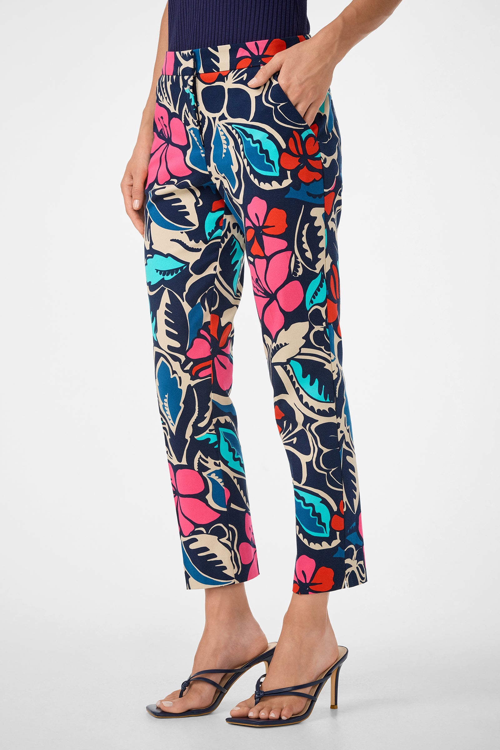 A person wears Moss 2 Pant—colorful, floral-print ankle pants with pink, red, blue, and cream patterns—paired with a navy top and black heeled sandals, standing with one hand in a pocket against a white background.