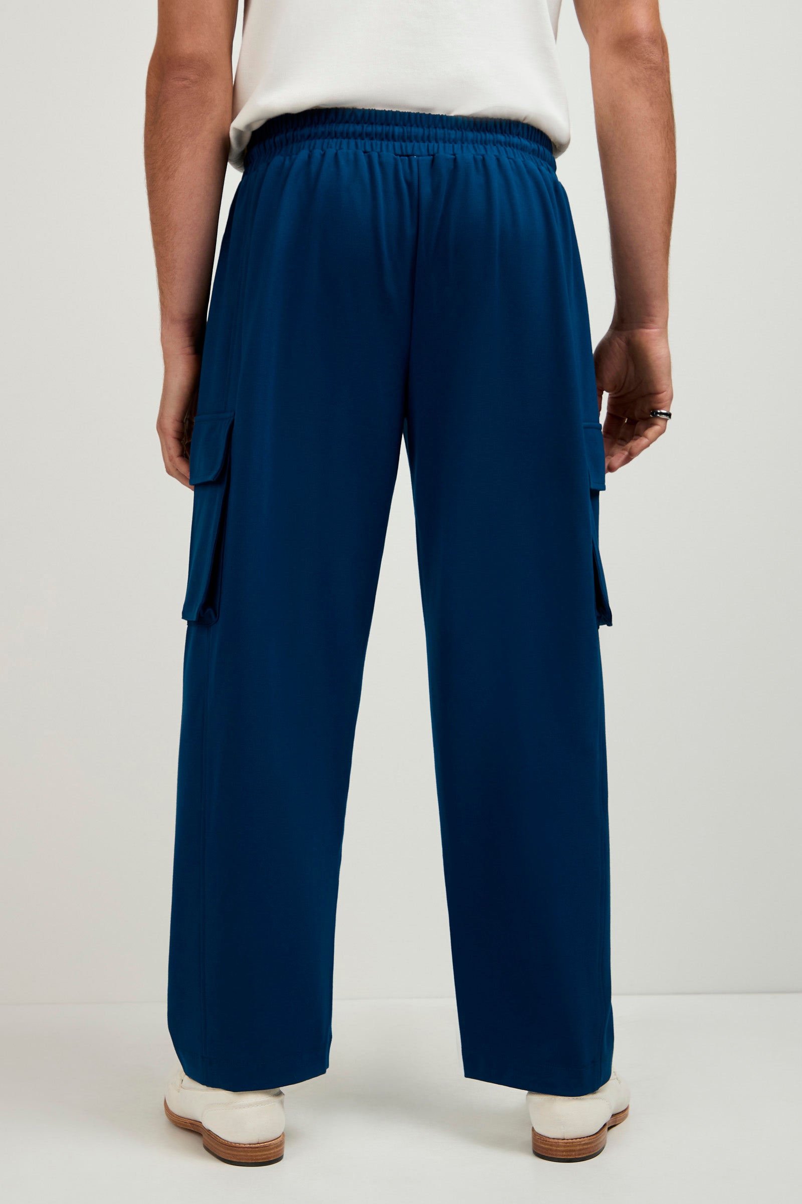 A person is shown from the back wearing the Lounge Cargo Pant in blue with cargo pockets, a white shirt tucked in, and white shoes, standing against a plain light background.