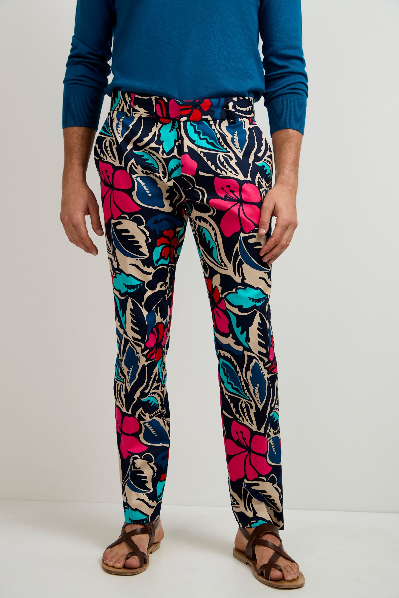 Wearing the Clyde Slim Trouser in Copacabana with bold floral patterns, paired with a blue long-sleeve shirt, matching belt, and brown sandals, a person stands against a plain background.