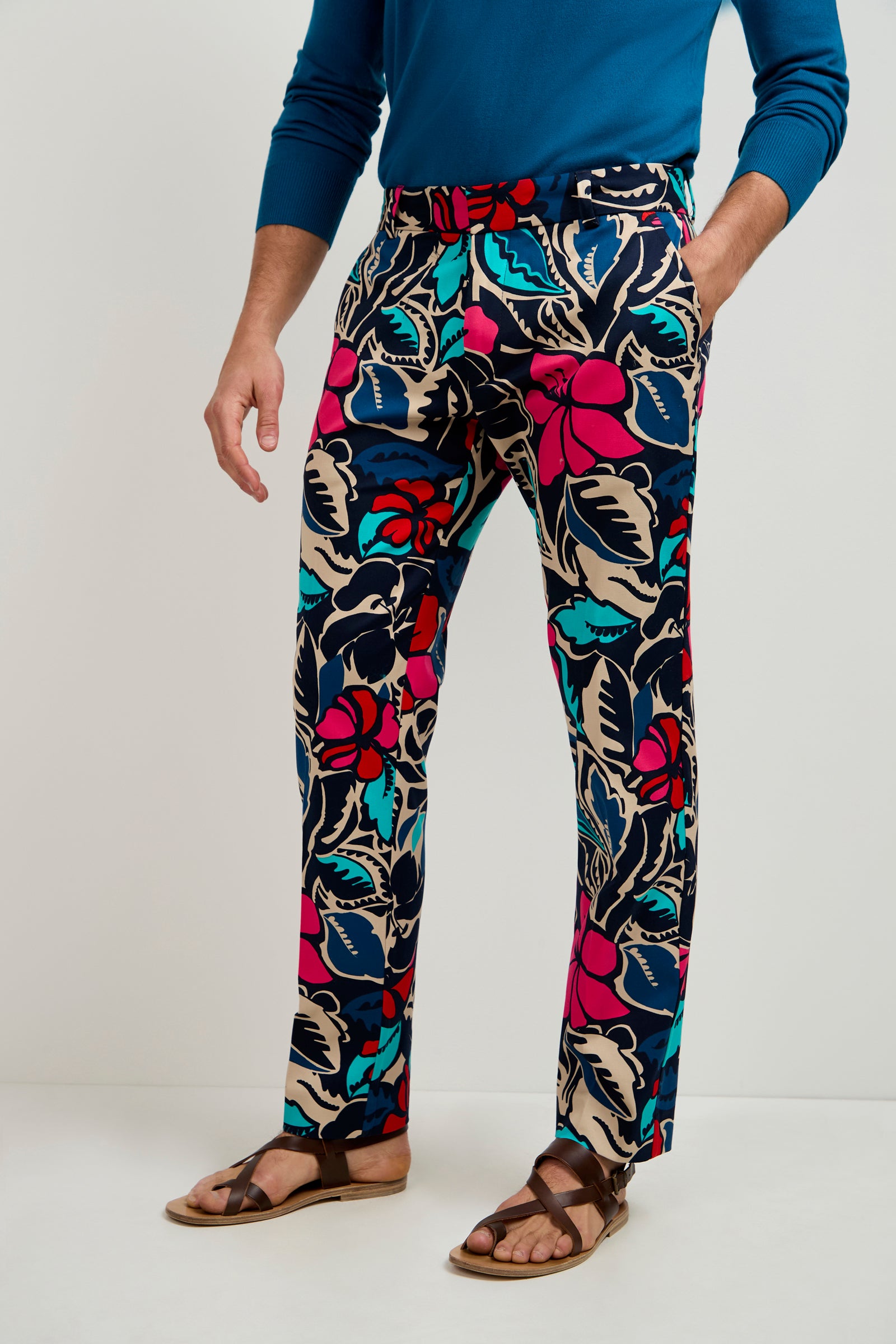 A person wearing a blue long-sleeve shirt, Clyde Slim Trouser - Copacabana featuring colorful floral prints, and brown sandals stands against a plain background.