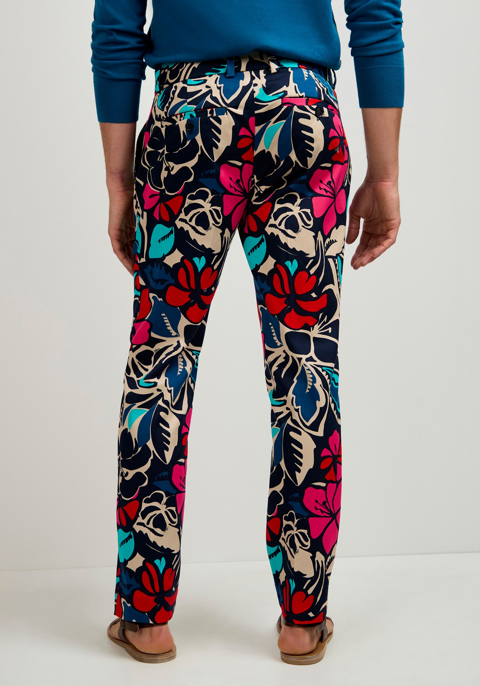 A person wearing the Clyde Slim Trouser - Copacabana, featuring a bold floral pattern in red, blue, teal, and beige, stands facing a white wall. They're also styled with a blue sweater and brown sandals.