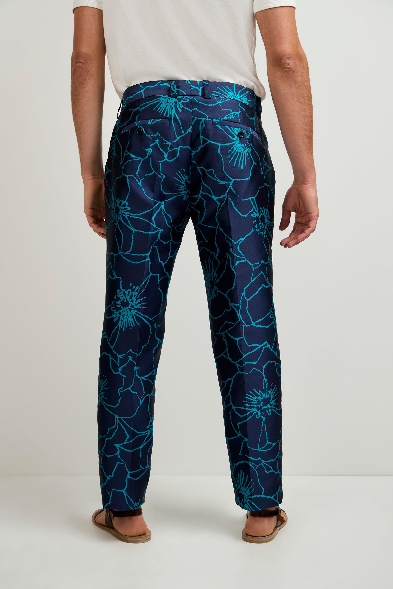 A person stands facing away, wearing the Clyde Slim Trouser - Jucara Jacquard, featuring a large bright blue floral pattern, paired with a white shirt and sandals.