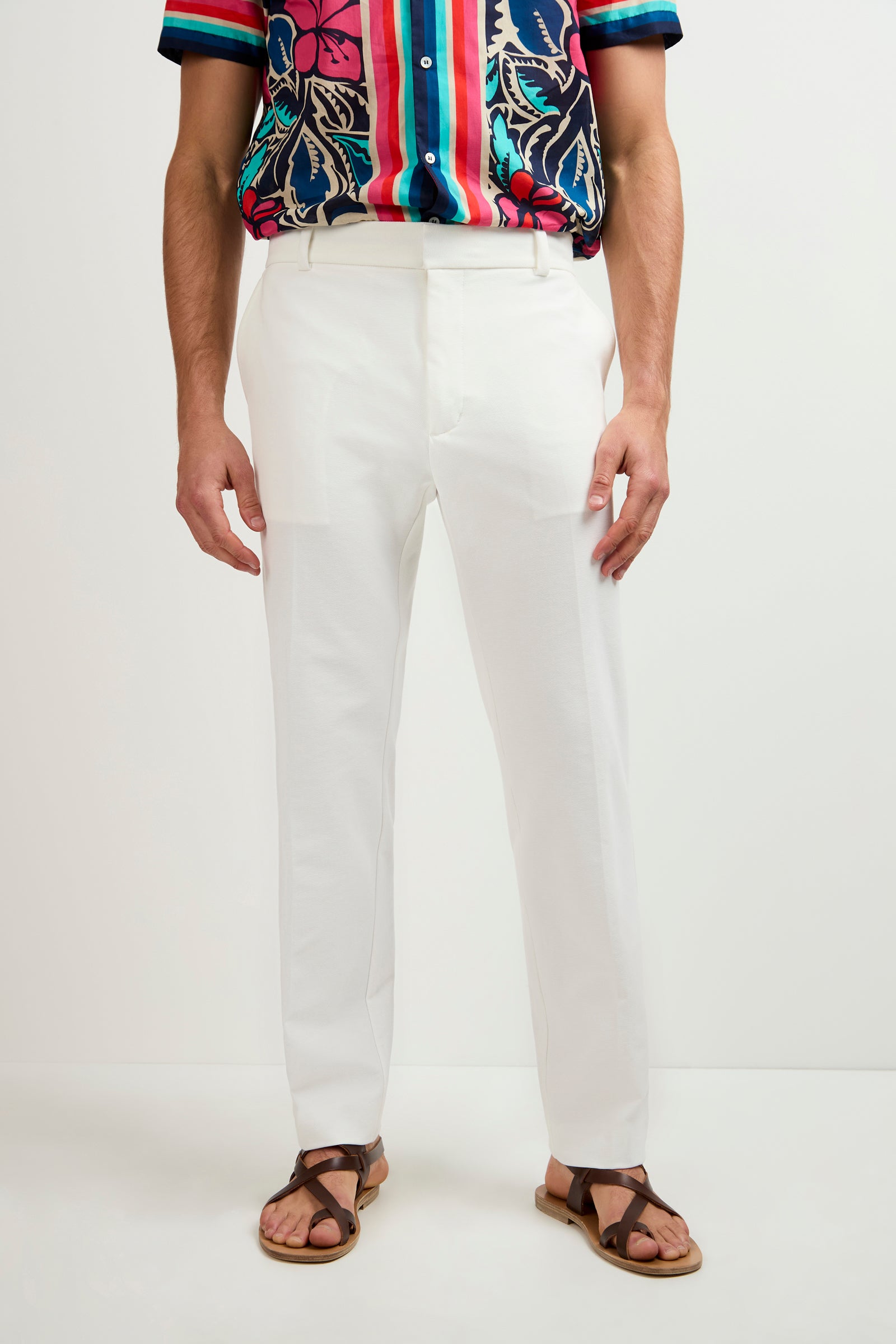 A person wearing the Clyde Slim Trouser - Stretch Piqué in Whitewash, paired with a colorful floral short-sleeve shirt and brown sandals, stands against a plain light background.