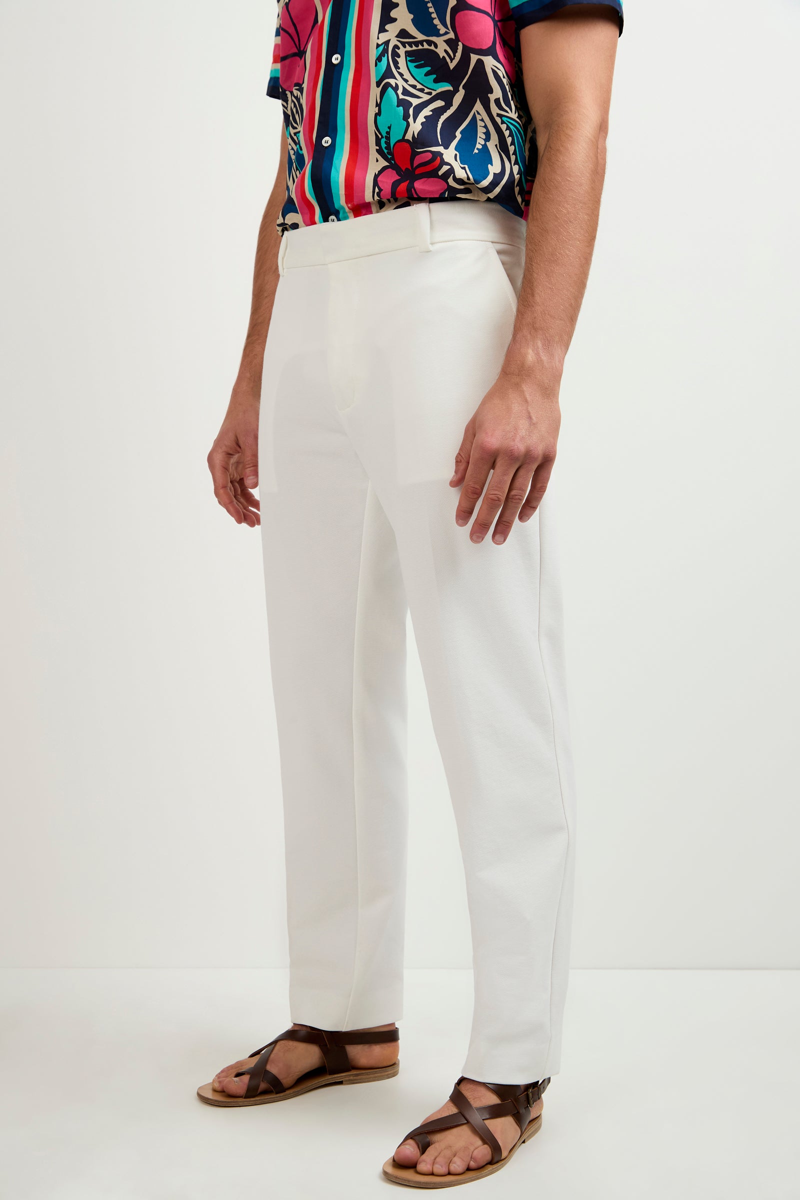 A person stands against a plain white background wearing the Clyde Slim Trouser - Stretch Piqué in white, paired with a colorful short-sleeved shirt and brown sandals. The person's face is not visible.