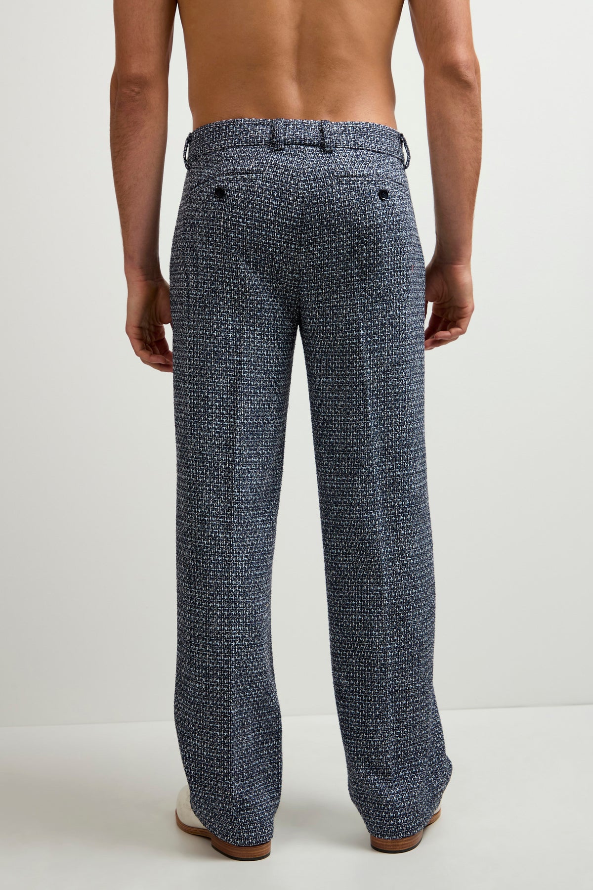 A shirtless man is shown from the back, barefoot in a plain white setting, wearing the Buckingham Tweed Trouser—loose-fitting dark blue pants with a textured look and small light-colored patterns.