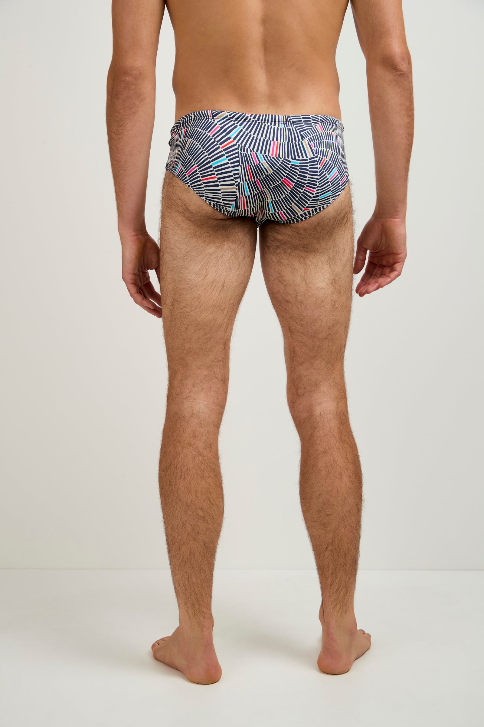 A man stands barefoot on a light floor, facing away from the camera, wearing the Bondi Swim Trunk - Java featuring a geometric pattern in black, white, and red. The background is plain and light-colored.