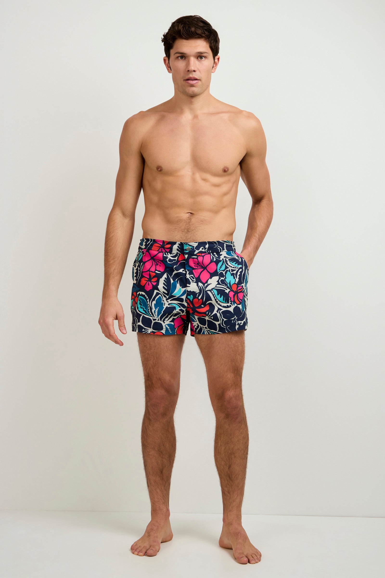 A man stands barefoot against a white wall, wearing the Chico Swim Trunk in Copacabana—a dark short featuring a vibrant floral print. He looks directly at the camera with a neutral expression.