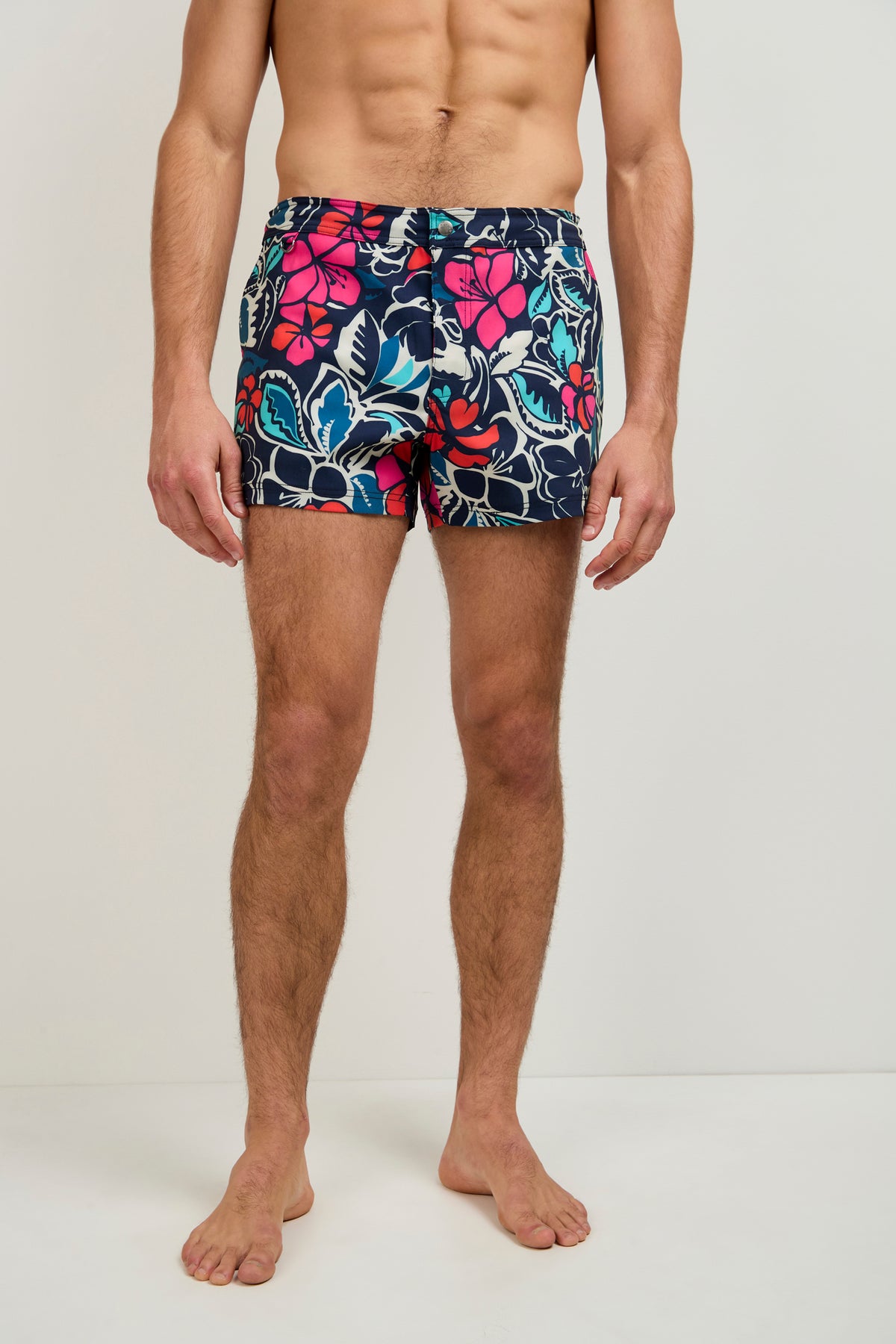 A man, barefoot against a plain background, models the Chico Swim Trunk - Copacabana in blue, pink, and white floral patterns. His upper body is bare and his face is not shown.