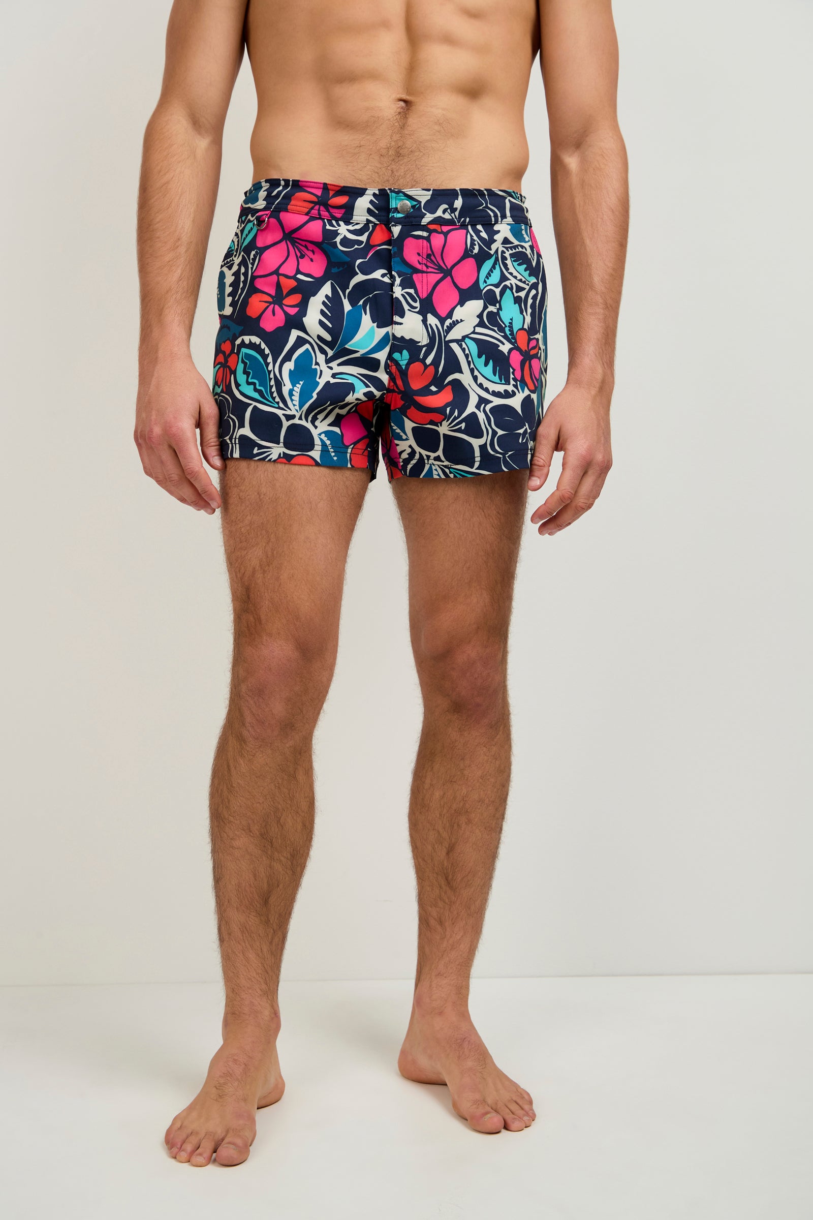 A man, barefoot against a plain background, models the Chico Swim Trunk - Copacabana in blue, pink, and white floral patterns. His upper body is bare and his face is not shown.