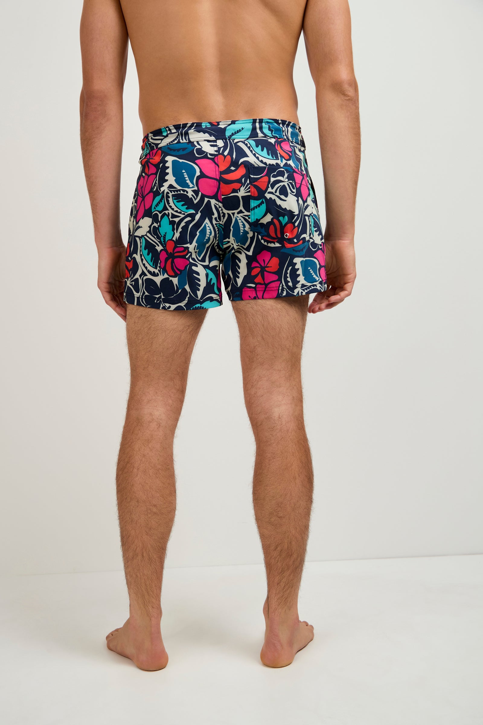 A man stands barefoot on a white floor, facing away from the camera, wearing the Chico Swim Trunk - Copacabana featuring a navy blue base with colorful red, pink, turquoise, and white floral patterns.