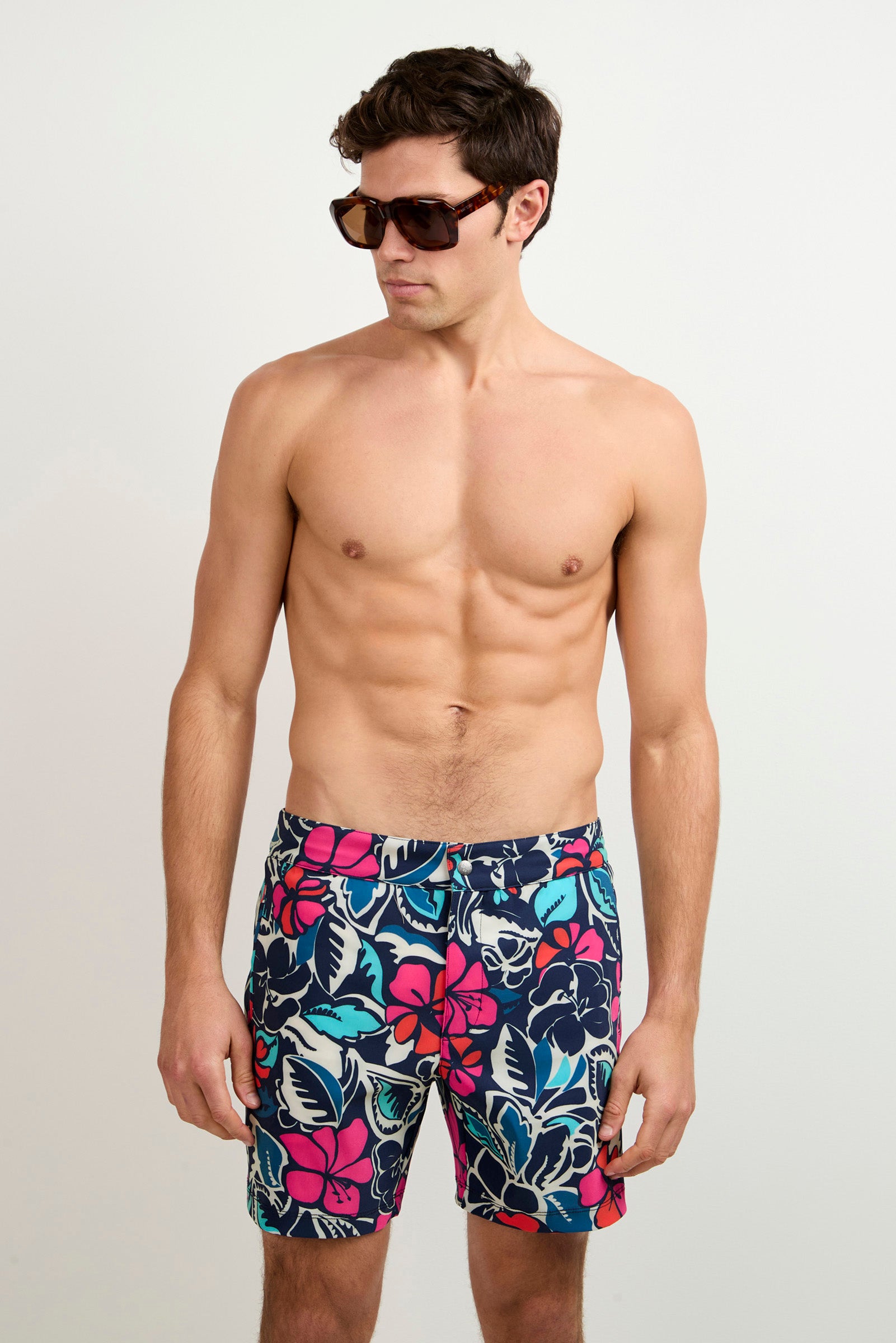 A man models the Honolulu Board Short - Copacabana, wearing sunglasses and colorful floral swim trunks, standing shirtless against a plain white background and looking slightly to the side.