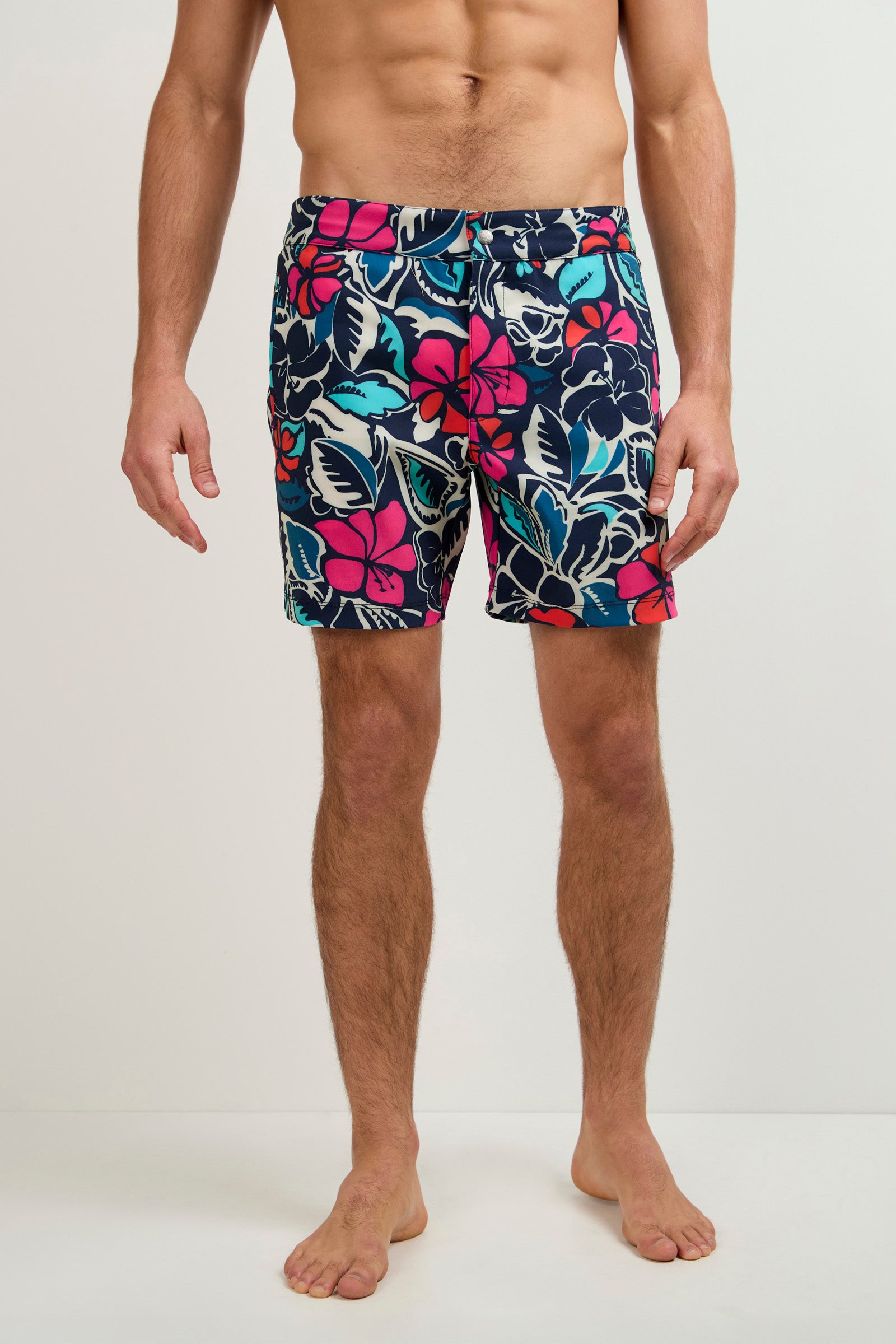 A man stands barefoot against a plain backdrop wearing the Honolulu Board Short - Copacabana, featuring colorful knee-length swim trunks with a bold pink, blue, and white floral print on a dark background. Image is cropped from shoulders to feet.