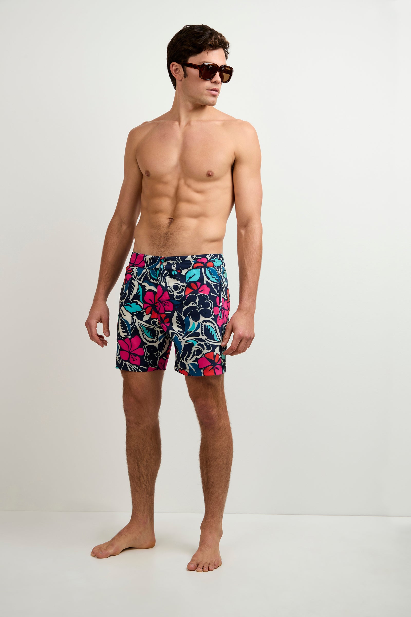 A man wearing the Honolulu Board Short - Copacabana stands barefoot against a plain white background, looking to his left while sporting sunglasses and colorful, floral-patterned swim trunks.