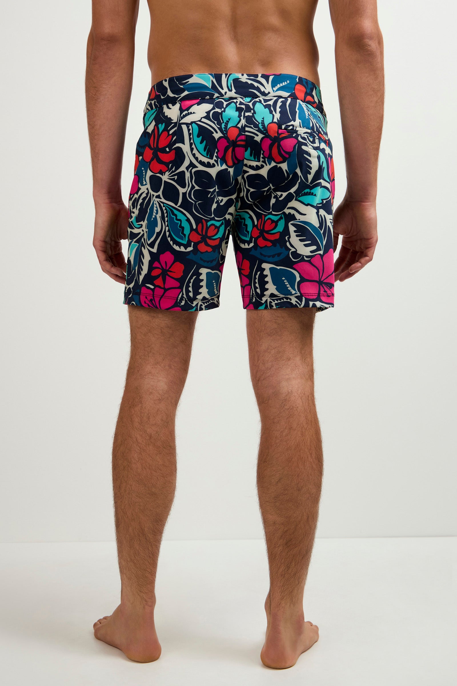 A man stands barefoot, facing away, wearing the Honolulu Board Short - Copacabana in a bold tropical floral print of red, blue, teal, and pink. The background is plain white.