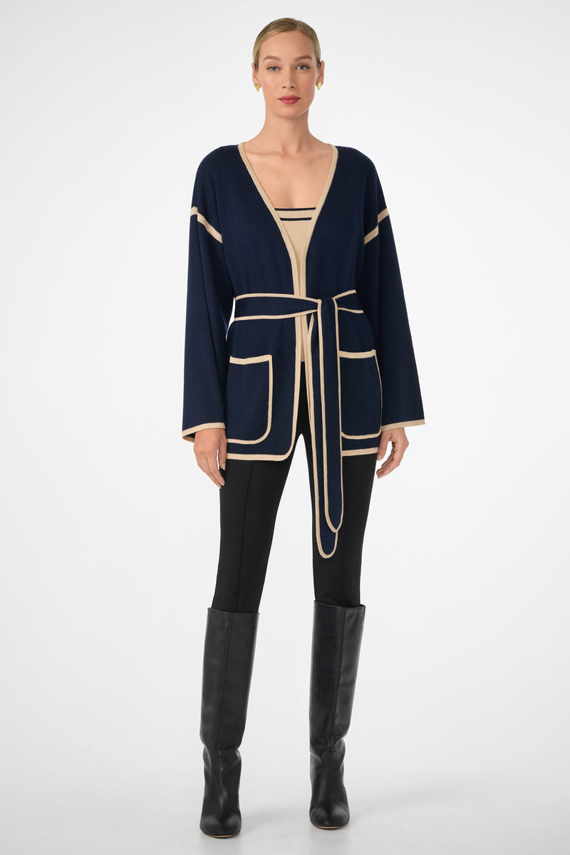 A woman faces forward against a white background, wearing the Wright Cardigan in navy blue with beige trim and a belt, paired with a beige top, black leggings, and black knee-high boots.