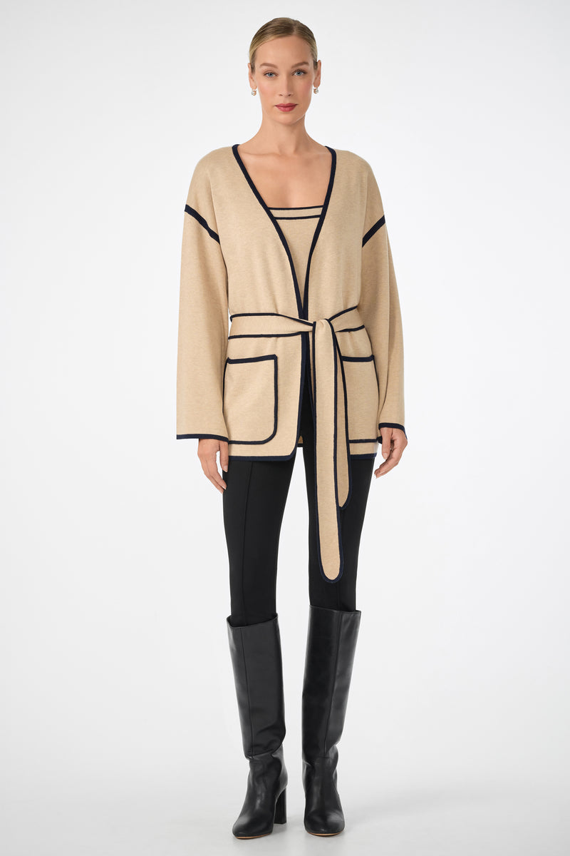 A woman stands facing forward in the Wright Wrap Cardigan—beige with black trim and a tied belt—paired with black pants and knee-high boots, set against a plain white background.