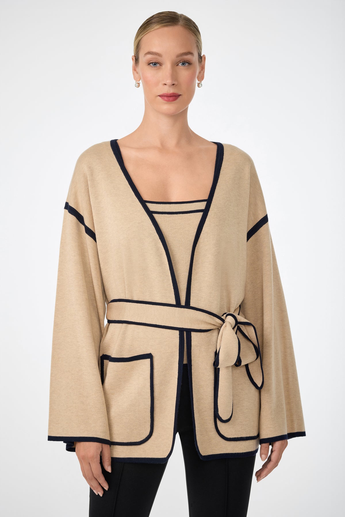 A woman wears the Wright Wrap Cardigan in beige with black trim, featuring wide sleeves, large front pockets, and a belted waist. She pairs it with a matching top and black pants, her hair pulled back, standing against a white background.